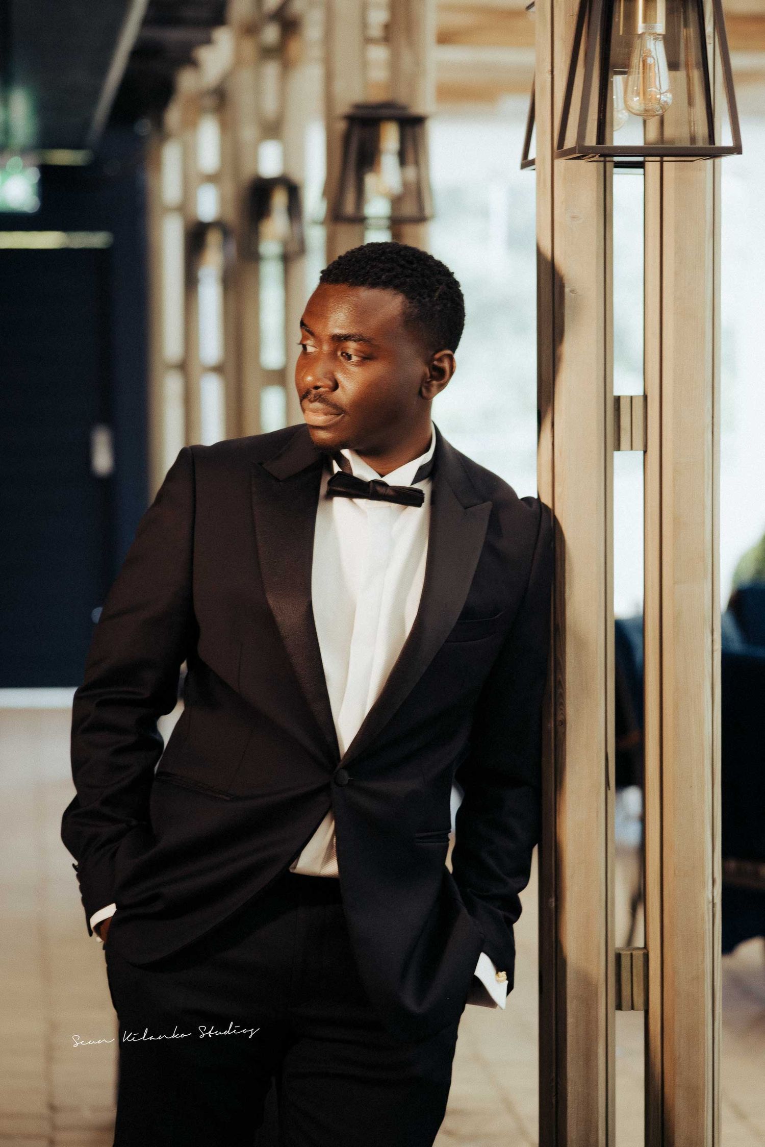 A groom in a classic black tuxedo poses confidently near an elegant light fixture.