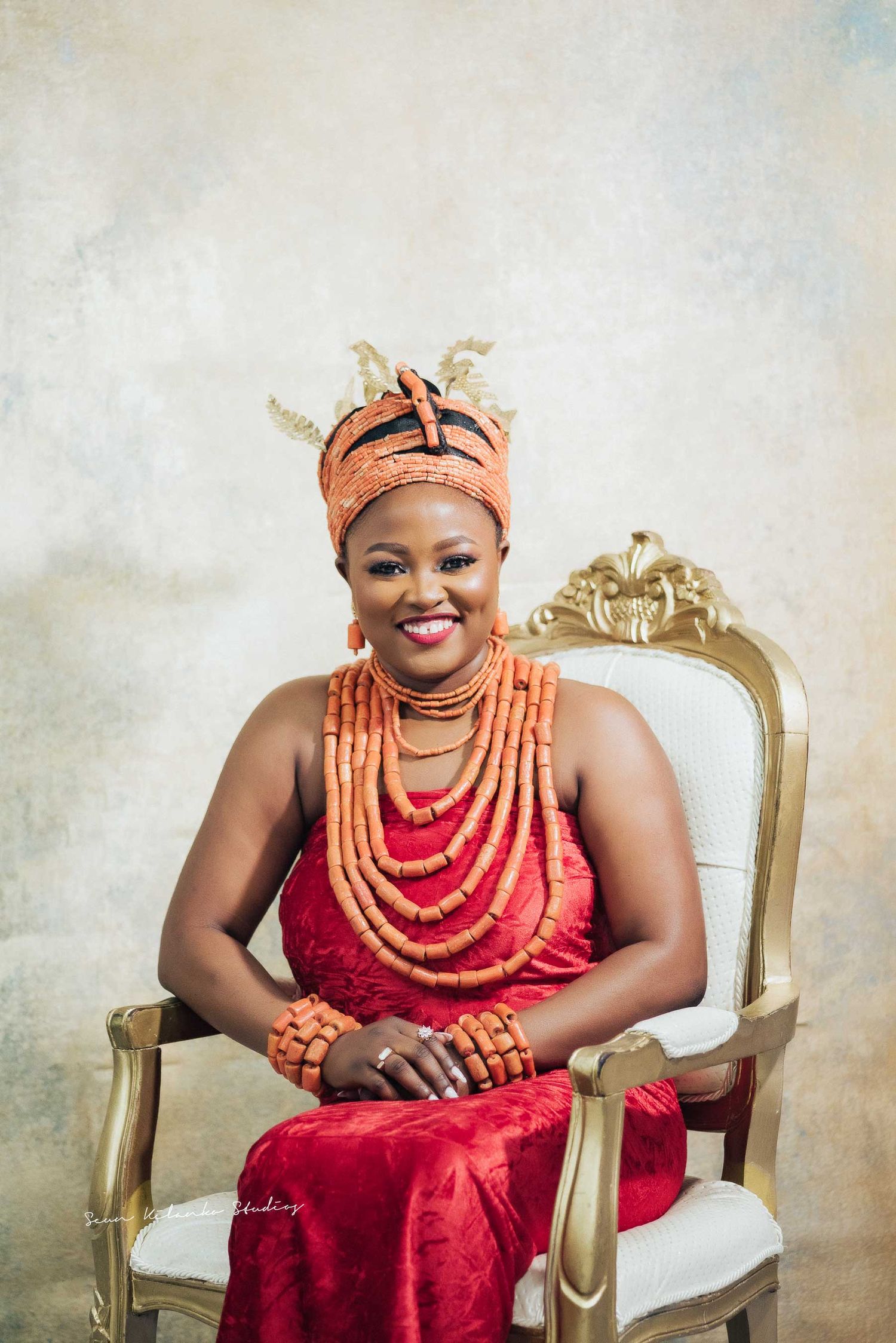 Elegant portrait in traditional coral beads and red dress seated on ornate gold chair.