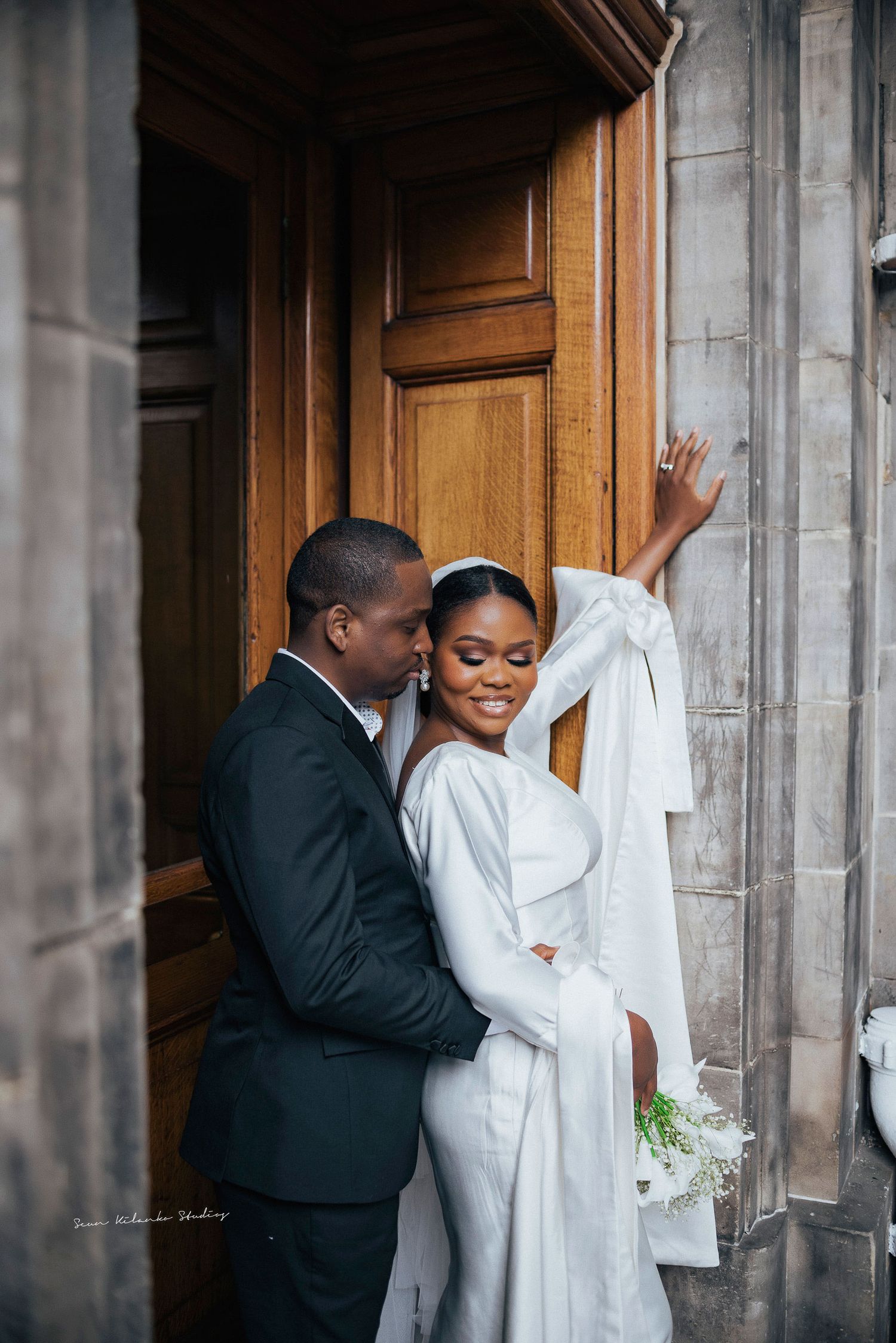 A tender wedding moment shared between newlyweds by a wooden door in an elegant architectural setting.