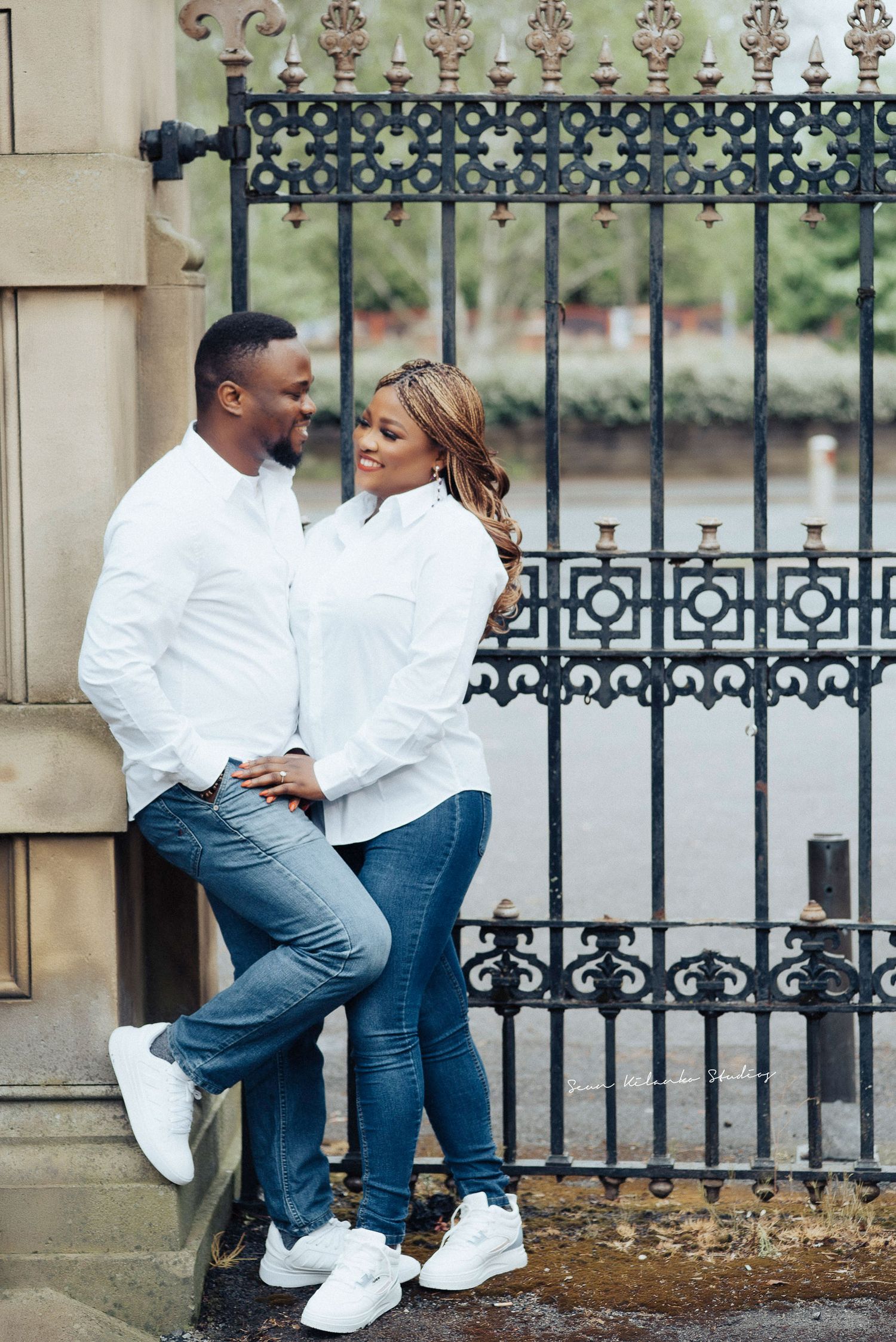 A couple in white shirts and jeans poses against an ornate iron gate.