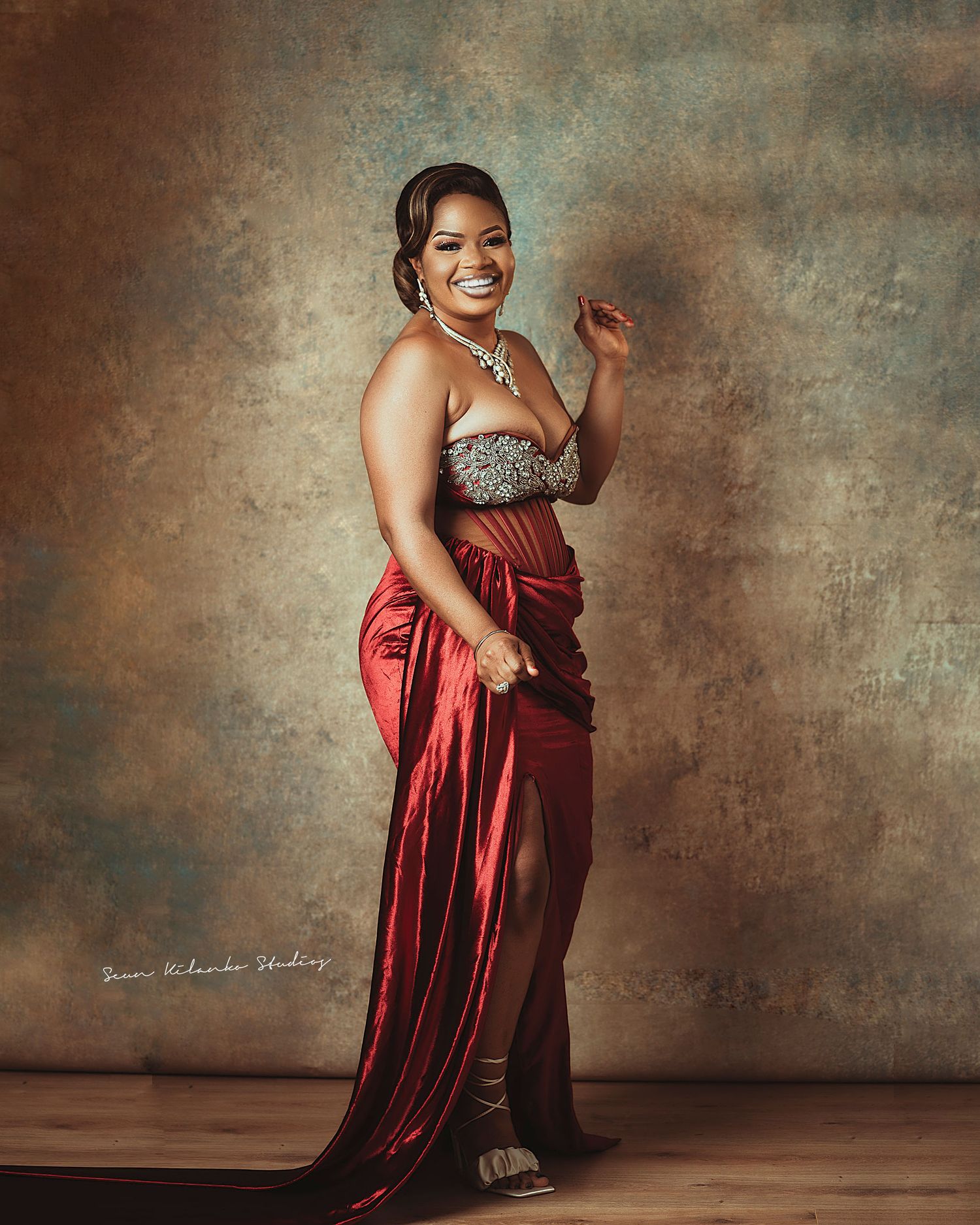 Professional studio portrait of a woman in a glamorous red silk dress and sparkly top posing against a textured background.
