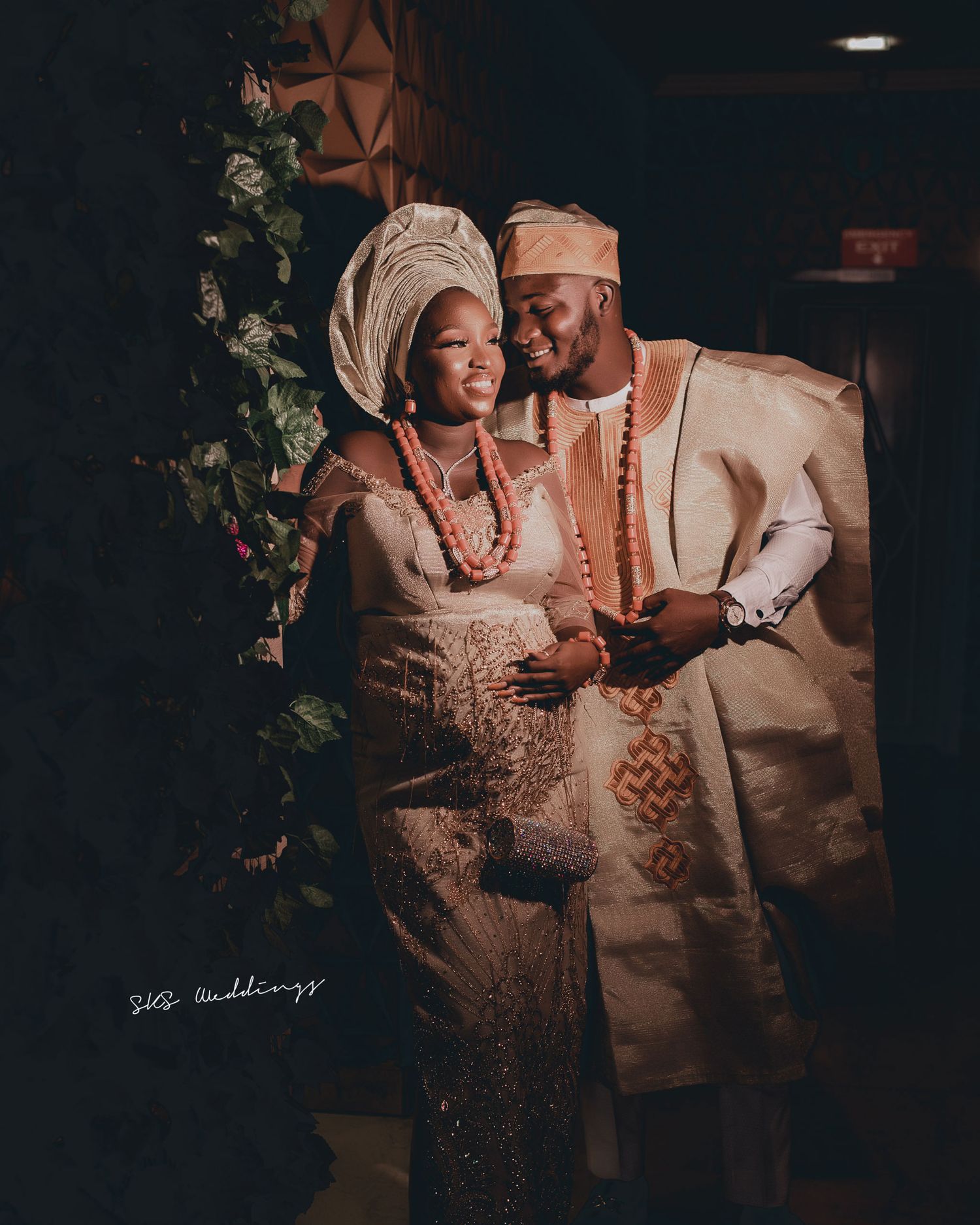 A couple in traditional Nigerian wedding attire share an intimate moment against a dark backdrop with decorative greenery.