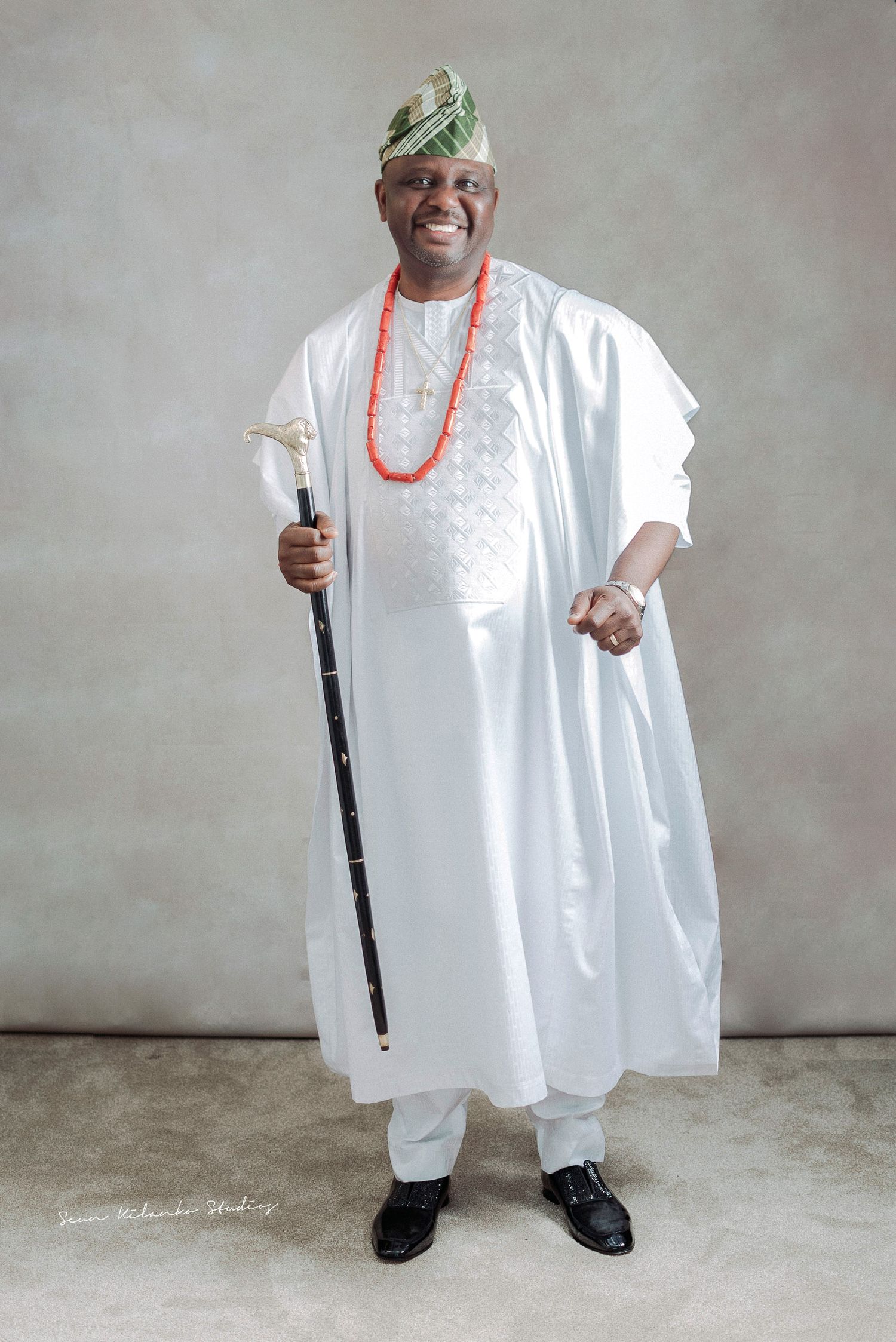 Traditional Nigerian attire with white agbada, green cap and coral beads holding ceremonial staff.