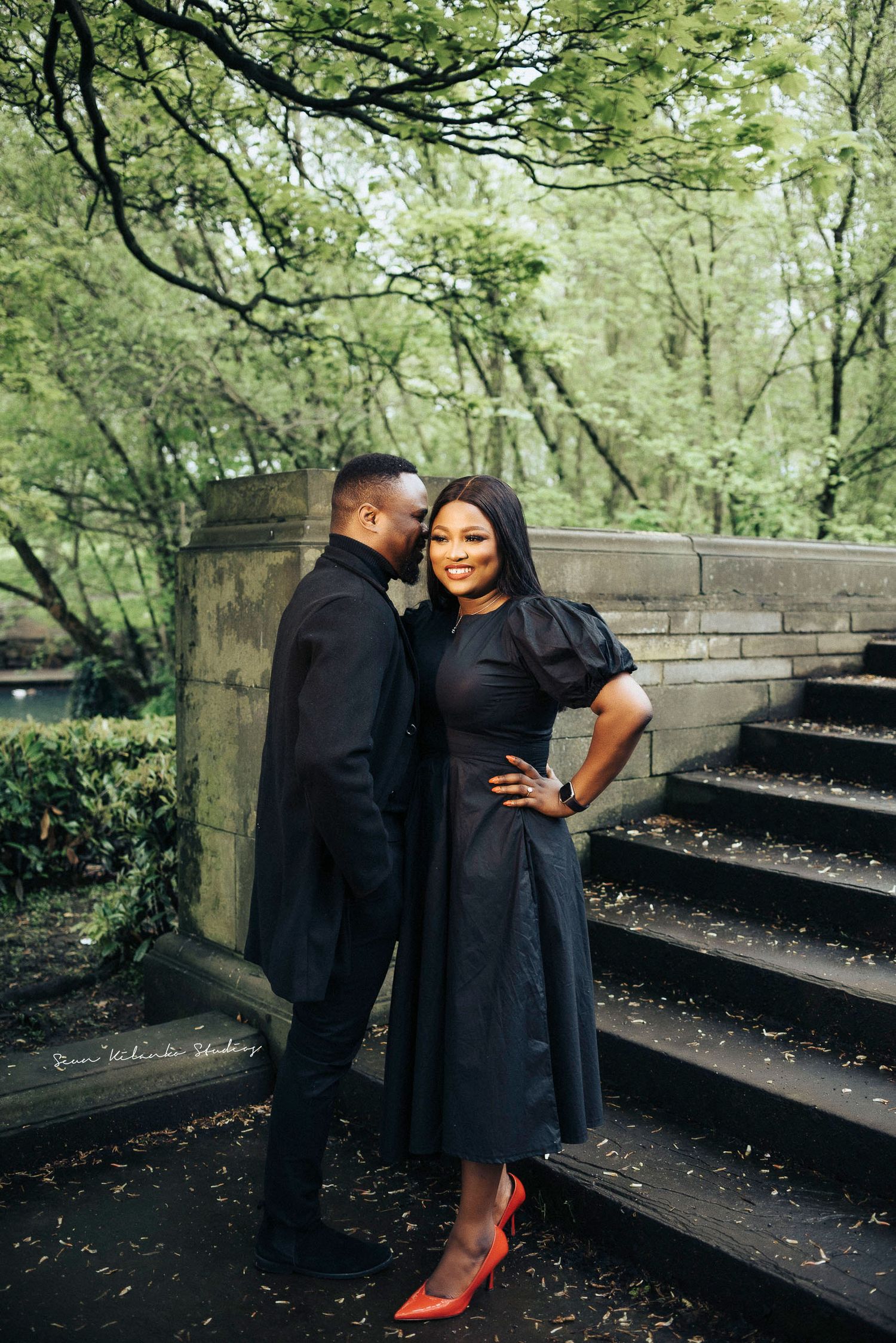 Romantic couple dressed in black poses together on stone stairs surrounded by greenery.