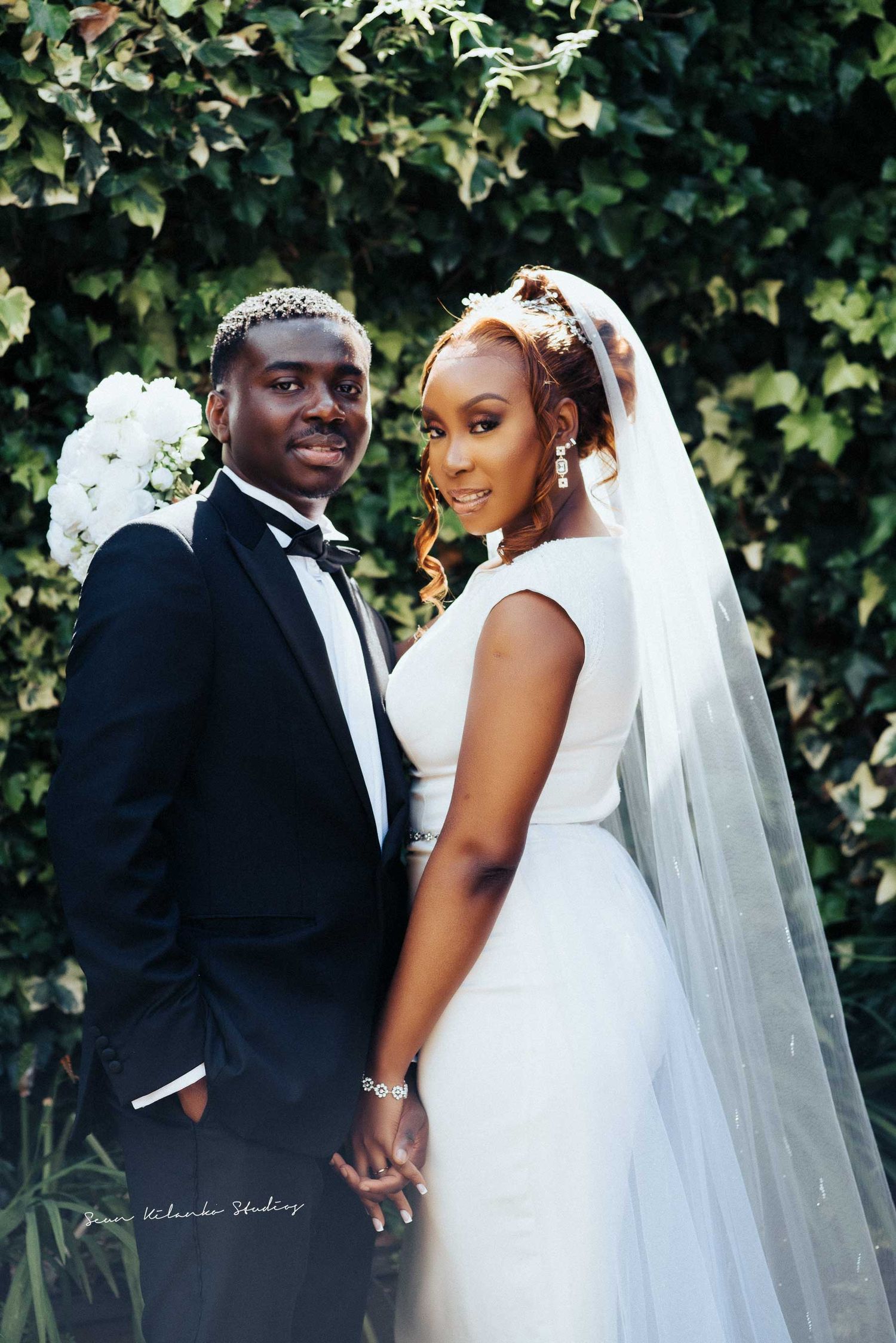 Elegant wedding couple poses in formal attire against lush garden greenery.