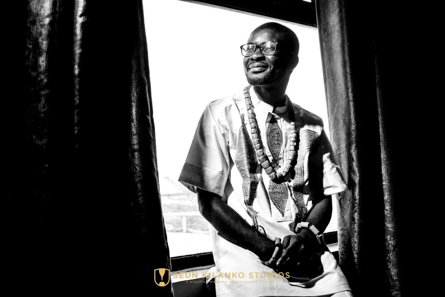 A black and white photograph shows someone smiling while wearing a light colored shirt and beaded necklace by a window.