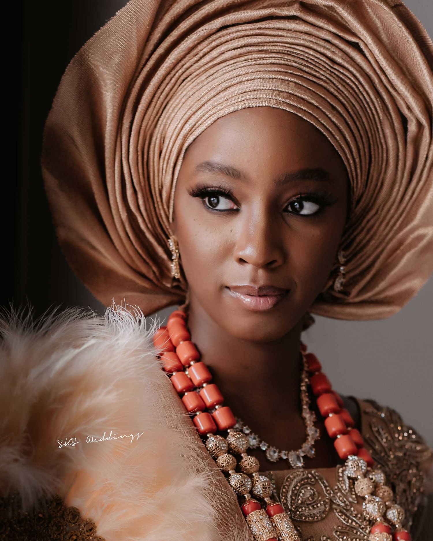 A portrait featuring coral beaded jewelry, an elegant brown gele headwrap, and white feather accents against a dark background.