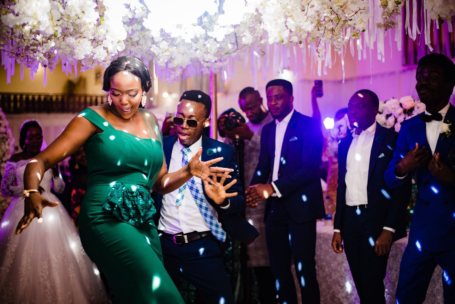 People dancing and celebrating on a dance floor at an elegant reception with purple lighting and floral decorations overhead.
