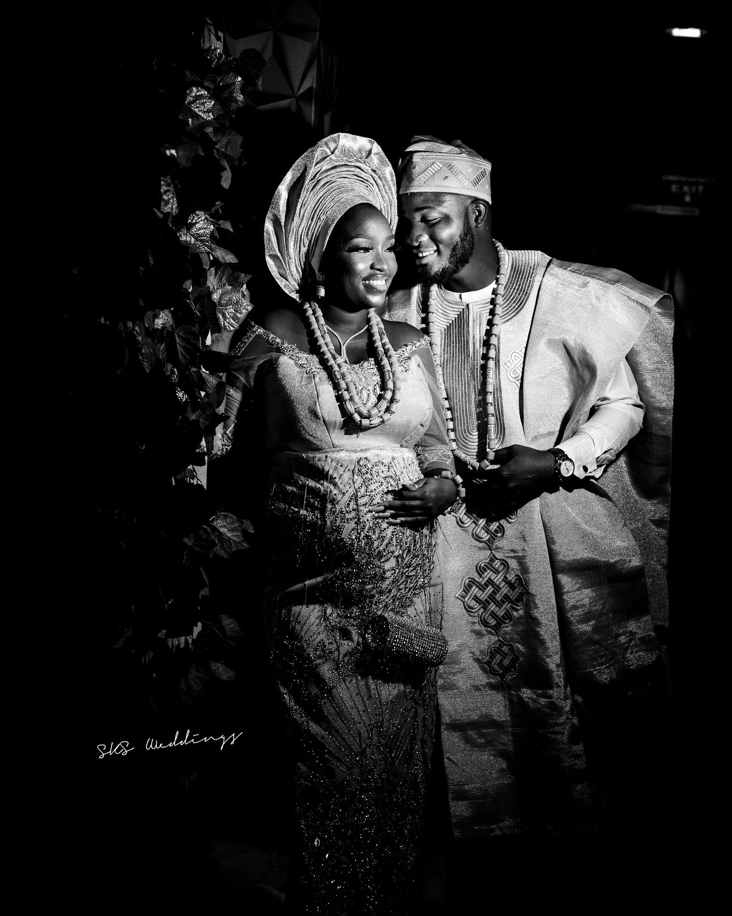 A black and white photograph shows two people in traditional Nigerian attire during what appears to be a special occasion.
