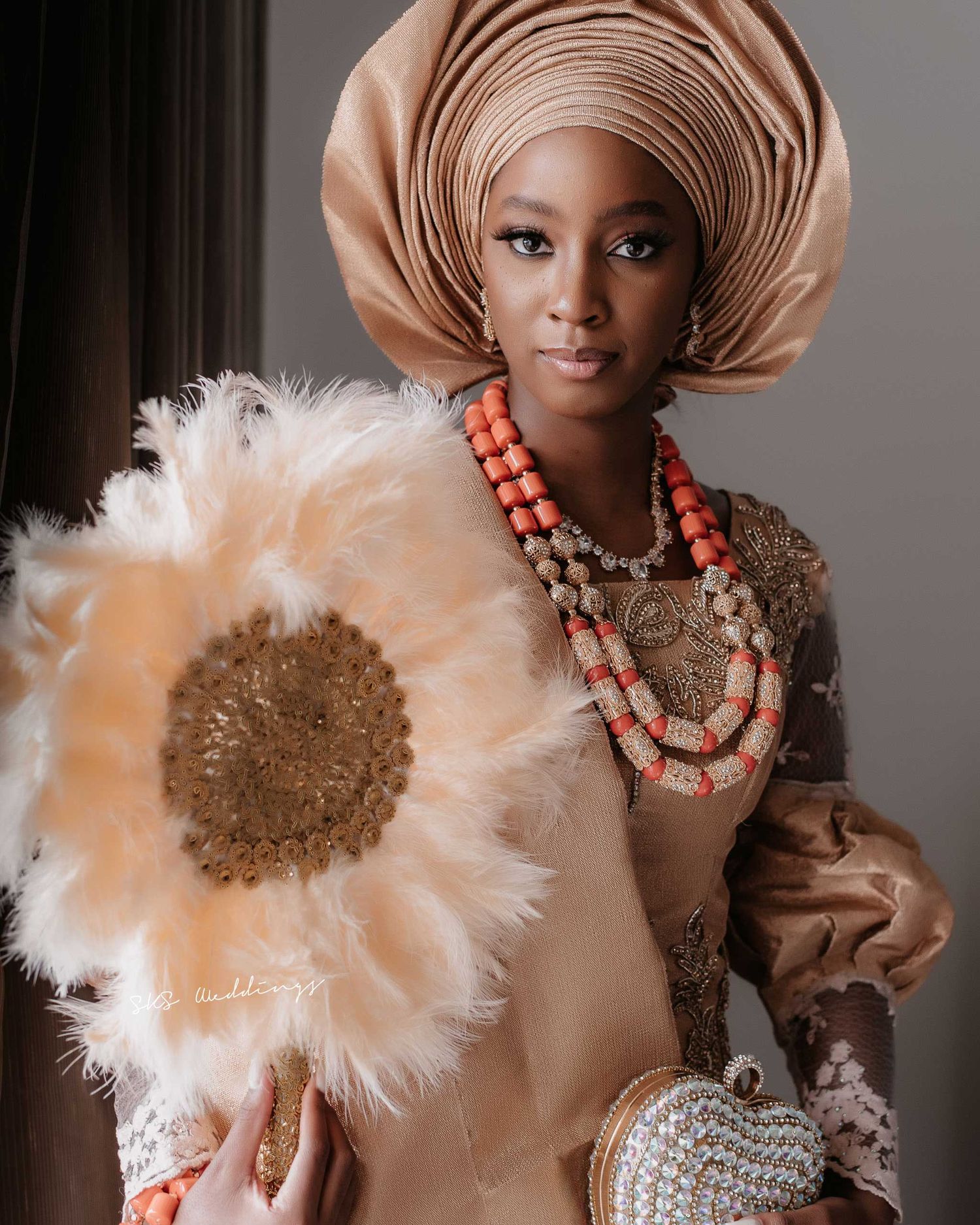 Traditional Nigerian wedding attire featuring a gold gele headpiece, coral beaded necklaces, and a large decorative white flower.
