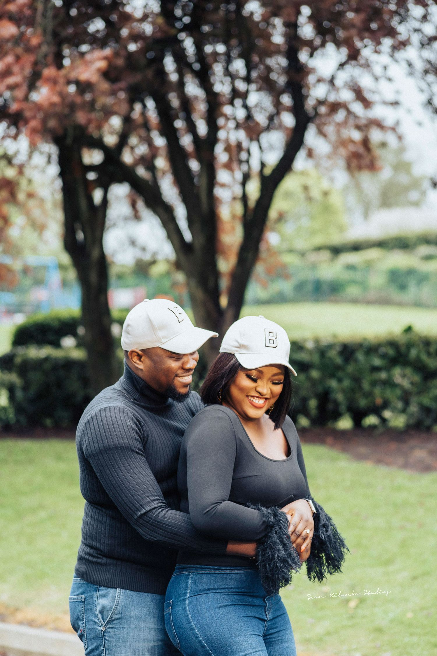 A couple in matching white baseball caps embraces in a park with autumn trees in the background.