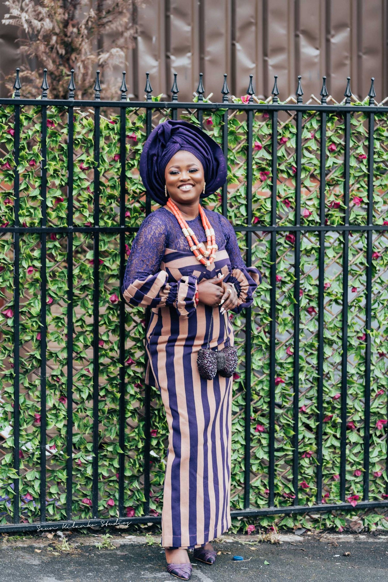 Nigerian fashion portrait against iron fence featuring purple gele and striped ankara dress.