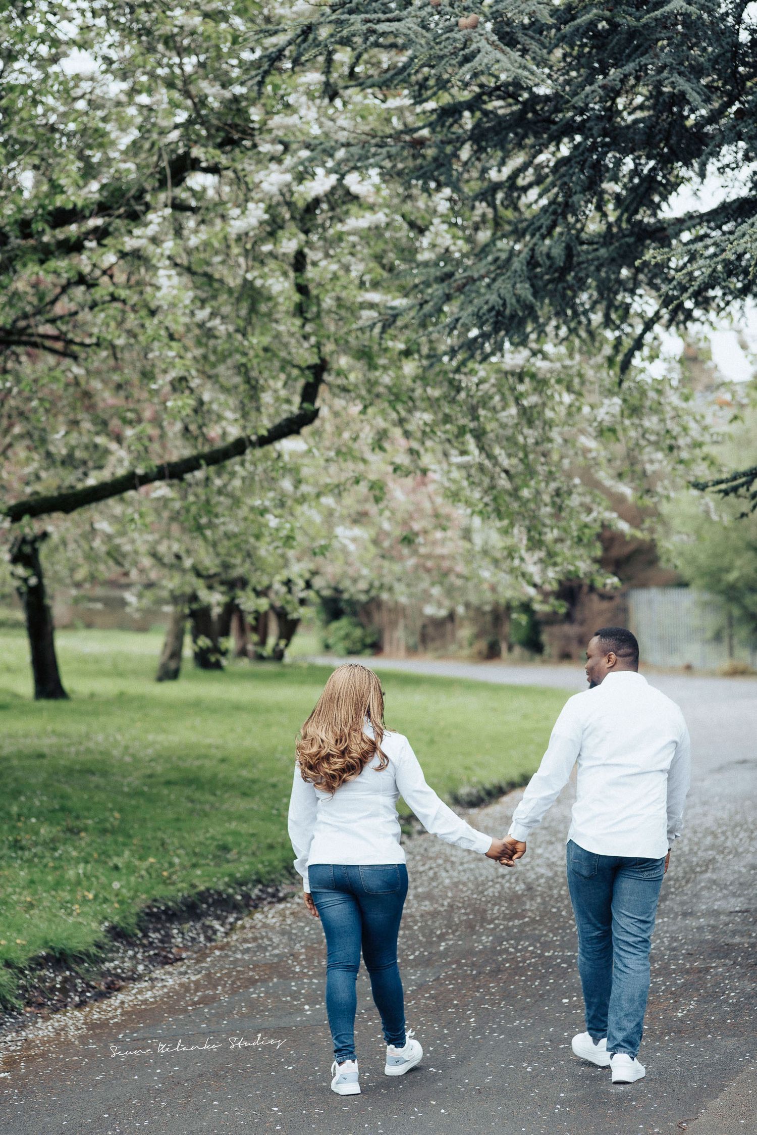 A couple walks hand in hand down a park path under blooming spring trees.