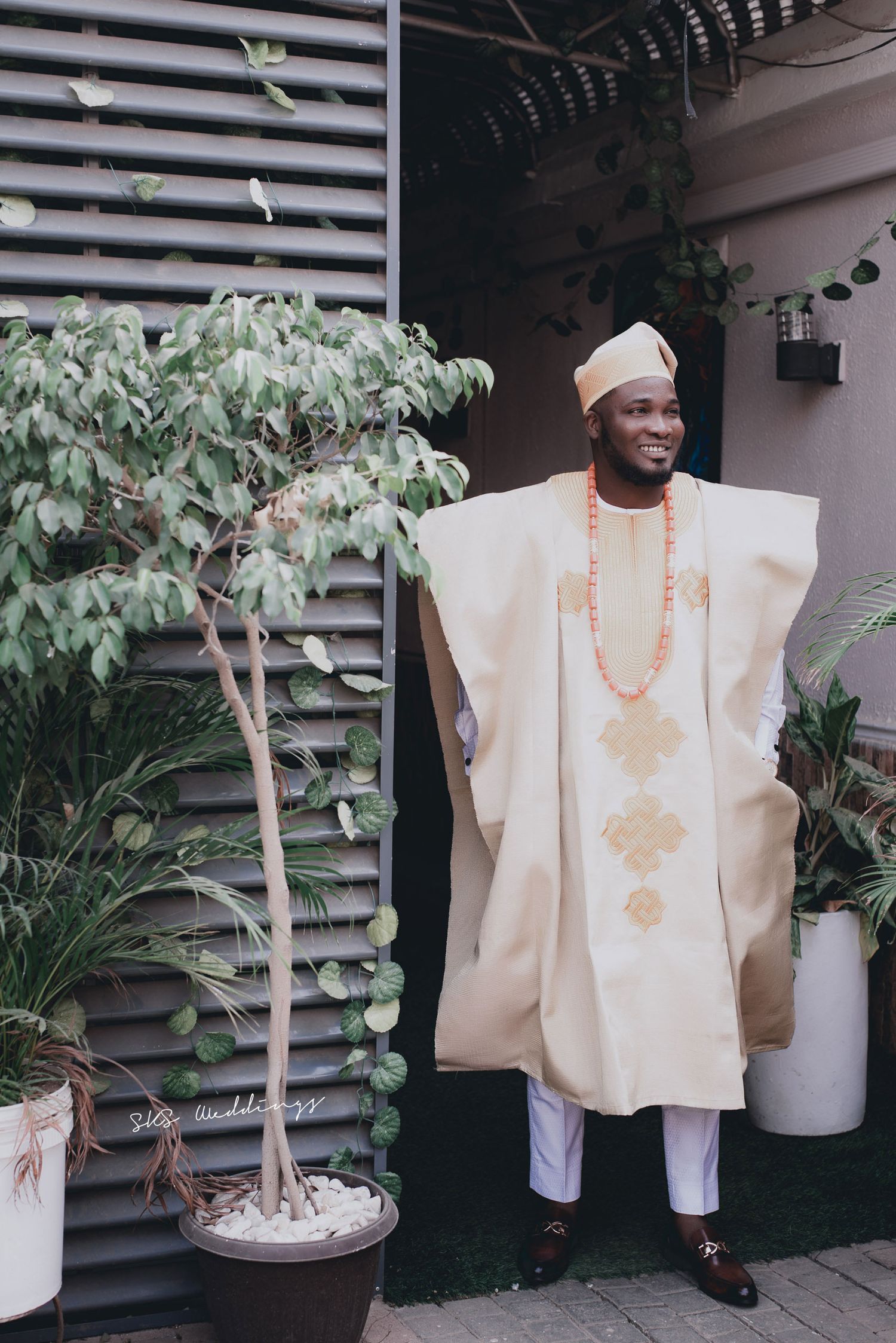 Traditional Nigerian agbada outfit in cream white with gold embroidery worn with beaded coral necklace by a small potted tree.