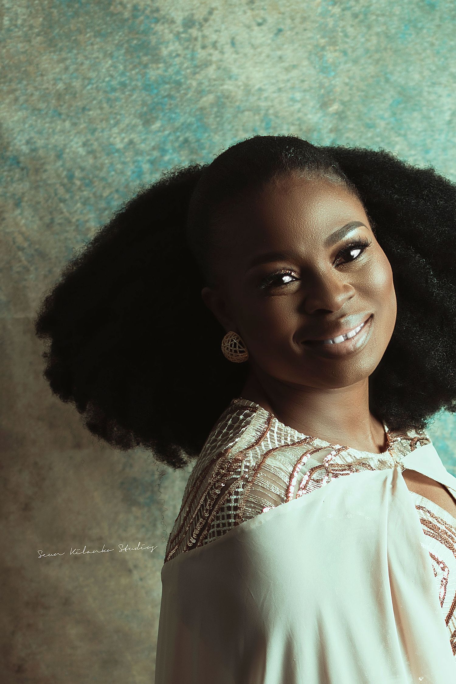 Portrait in cream lace dress with natural hair styled against turquoise background.