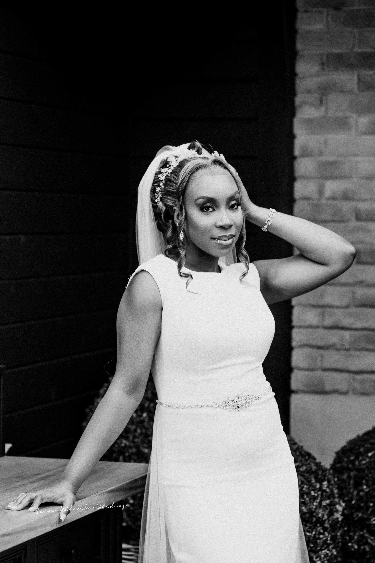 Black and white portrait of a bride in a fitted gown with veil adjusting her hair.