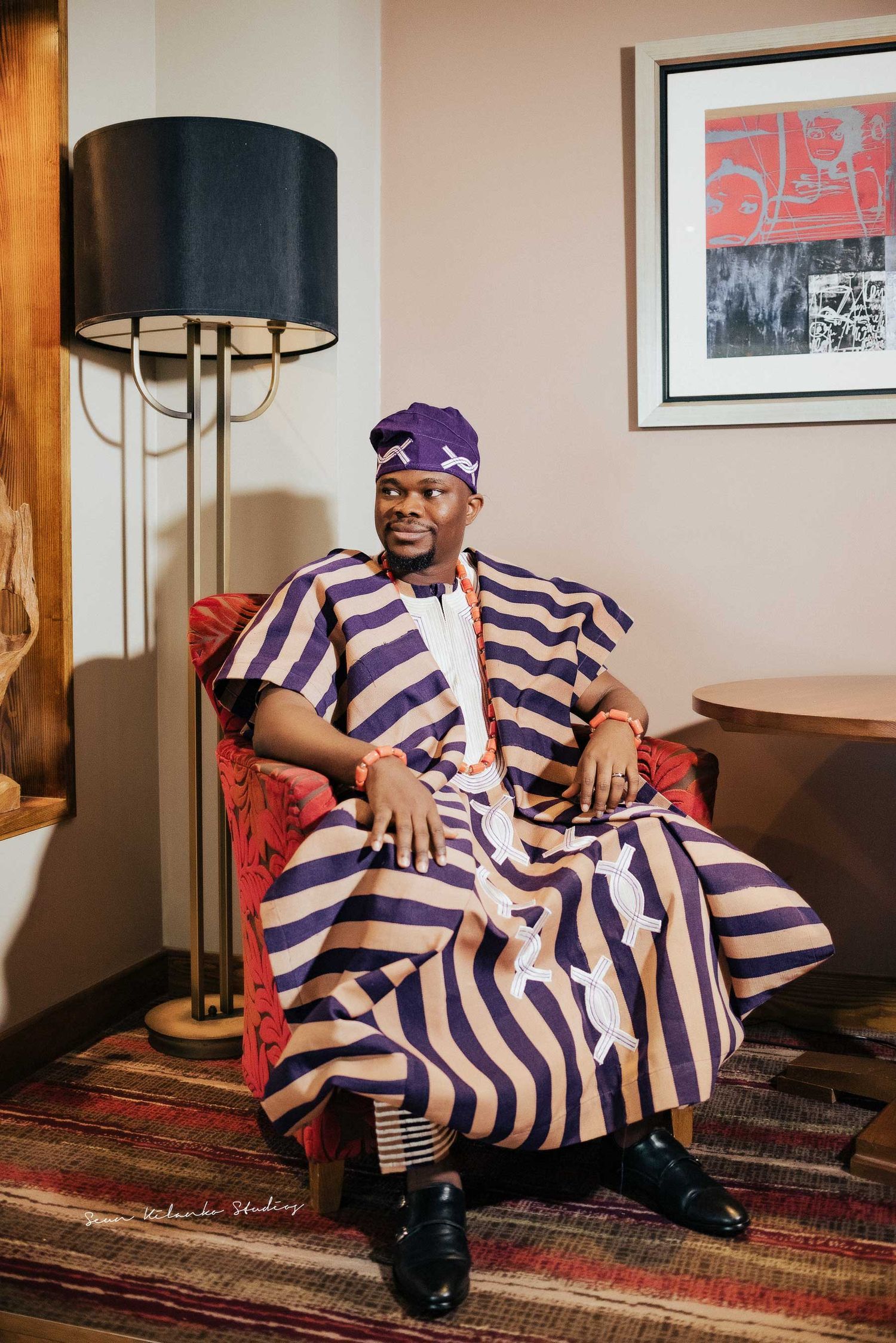 Traditional Nigerian attire showcased as a person sits in a striped agbada with purple headwrap on red chair.