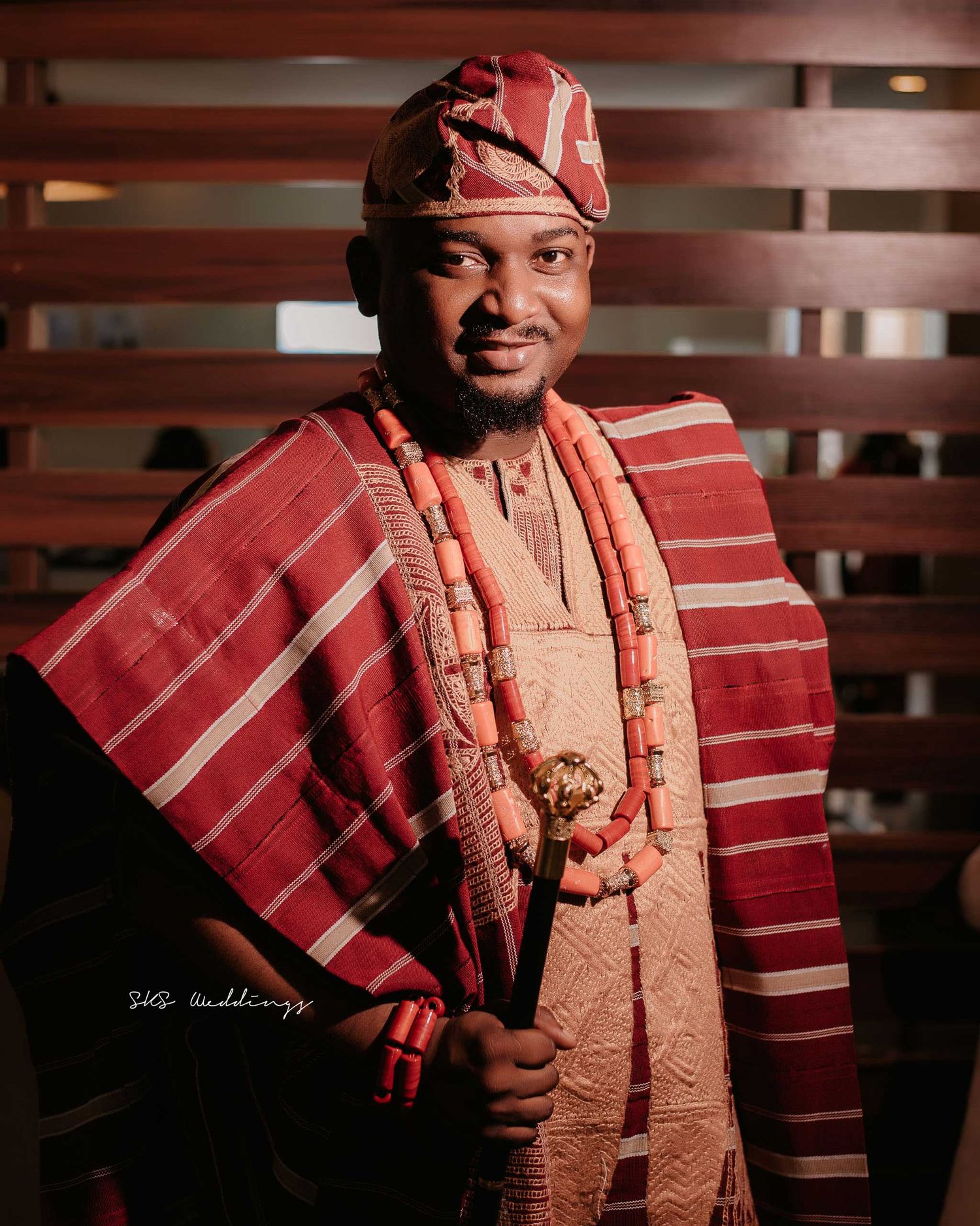 A person wearing traditional red and peach colored Nigerian attire with agbada and beaded accessories poses against wood paneling.