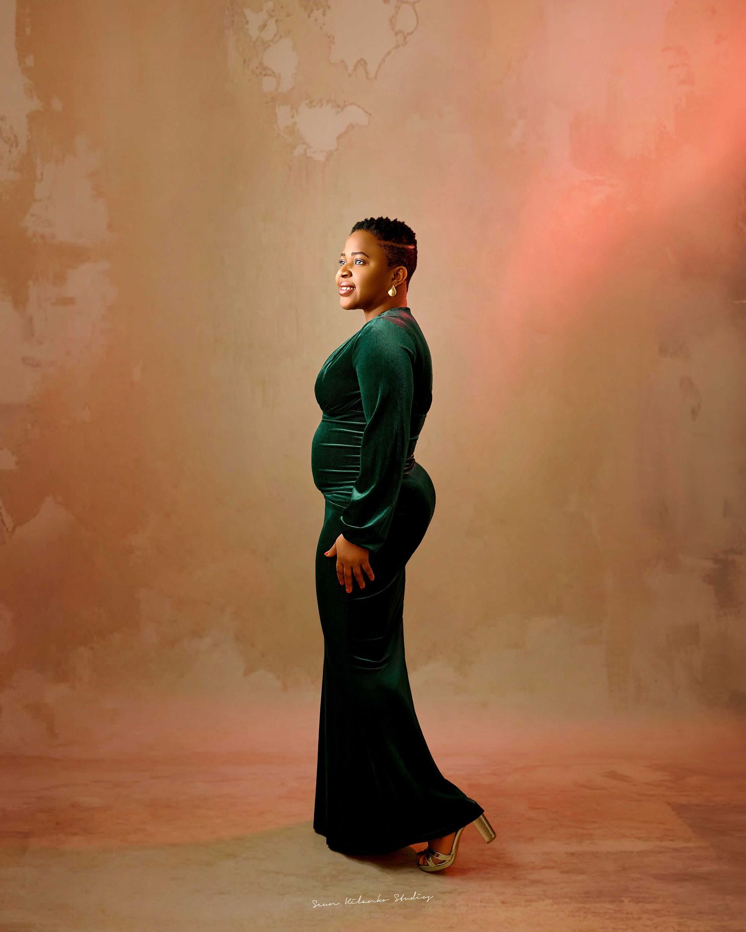 Pregnant woman in elegant green velvet gown poses sideways in studio maternity portrait against warm beige backdrop.