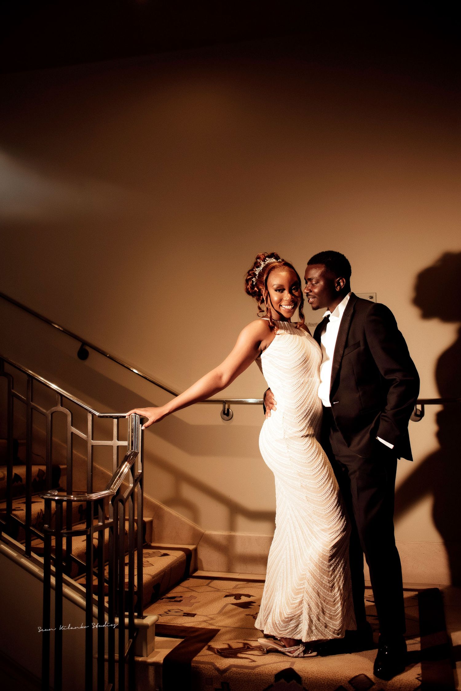Elegant couple in formal attire posing on a staircase, woman in white gown and man in black tuxedo.