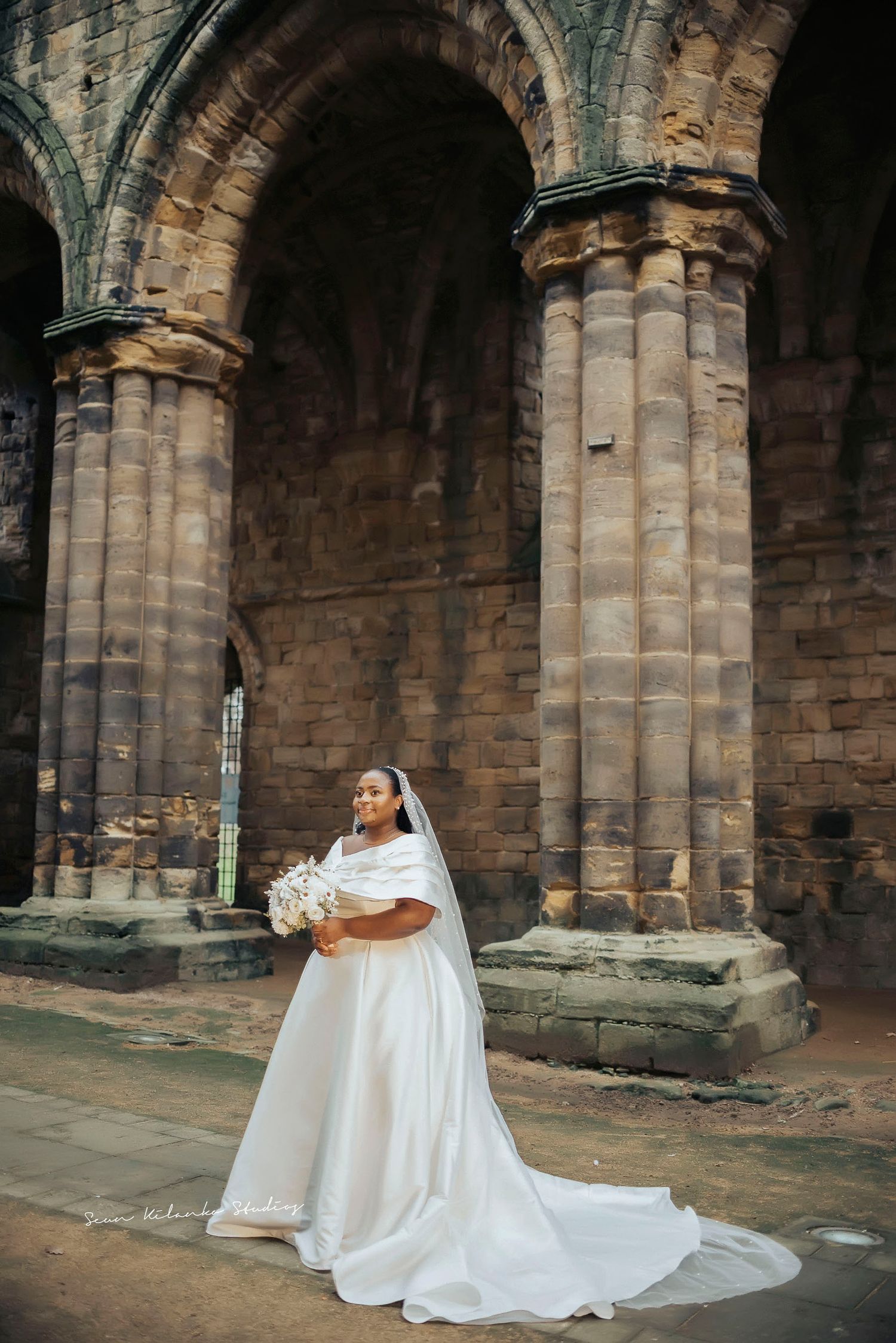A bride in a beautiful white gown poses alone under gothic cathedral architecture.