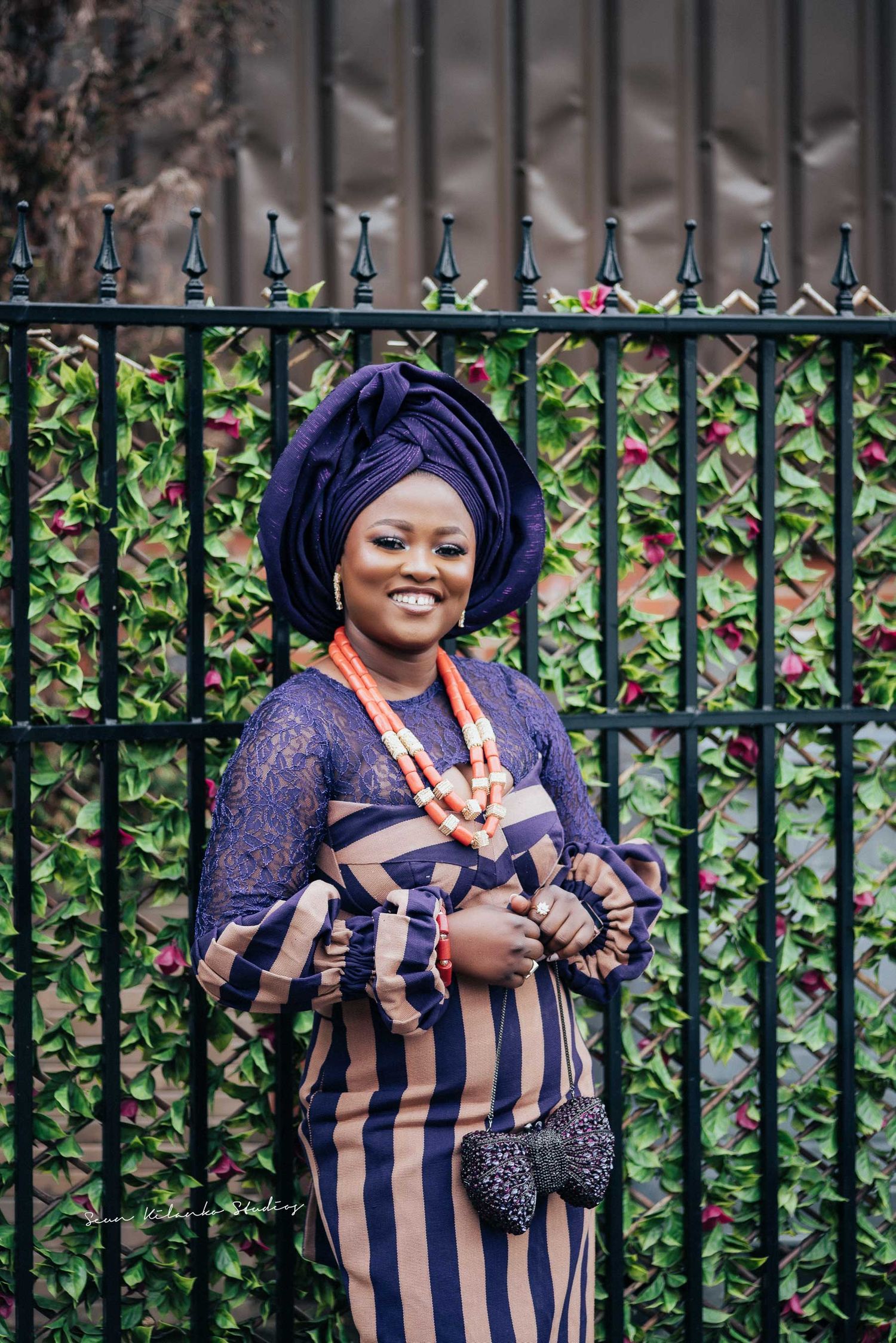 Traditional African fashion portrait featuring navy gele headwrap and striped dress against ornate iron fence.