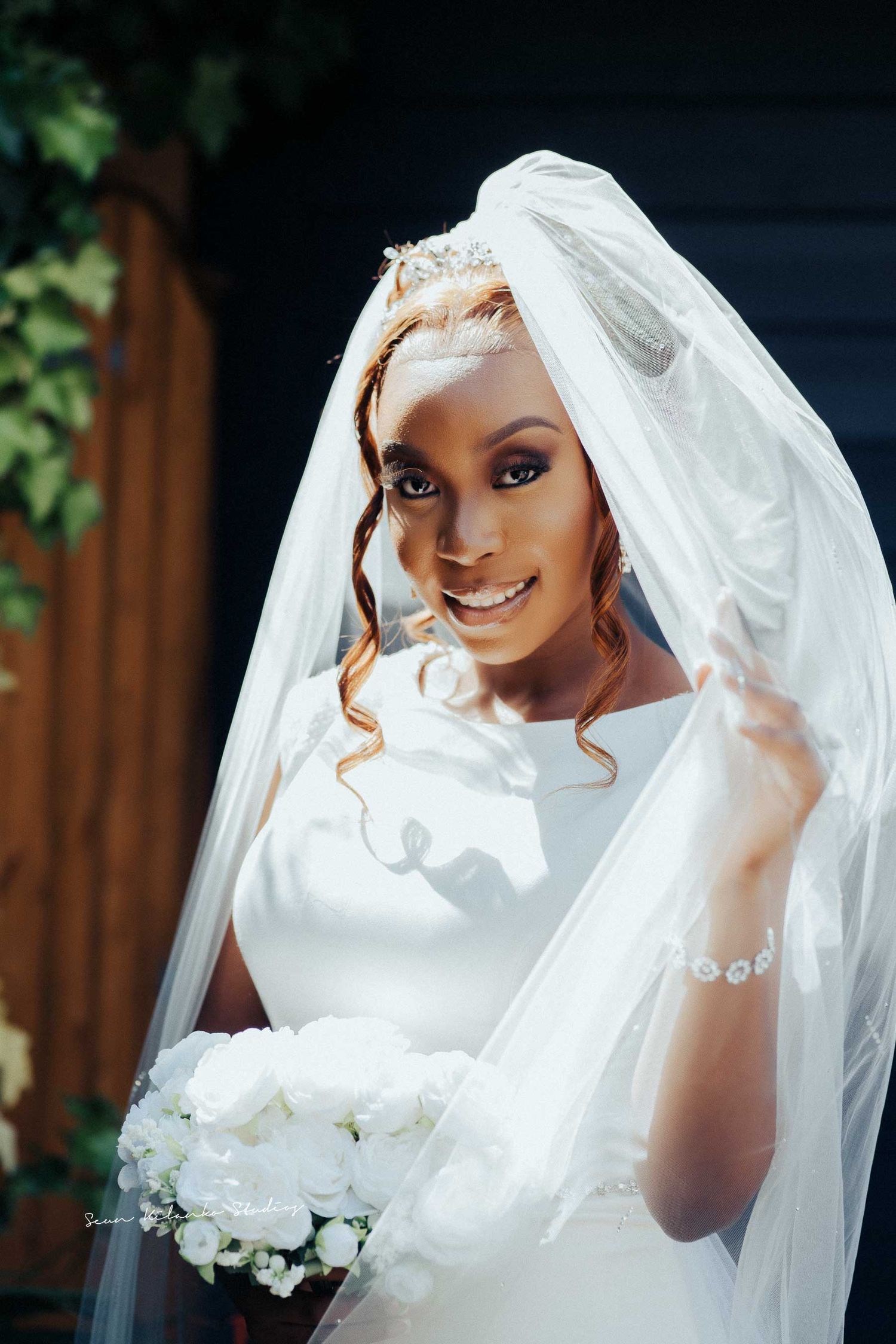 Radiant bridal portrait with flowing veil and white bouquet against wooden backdrop.
