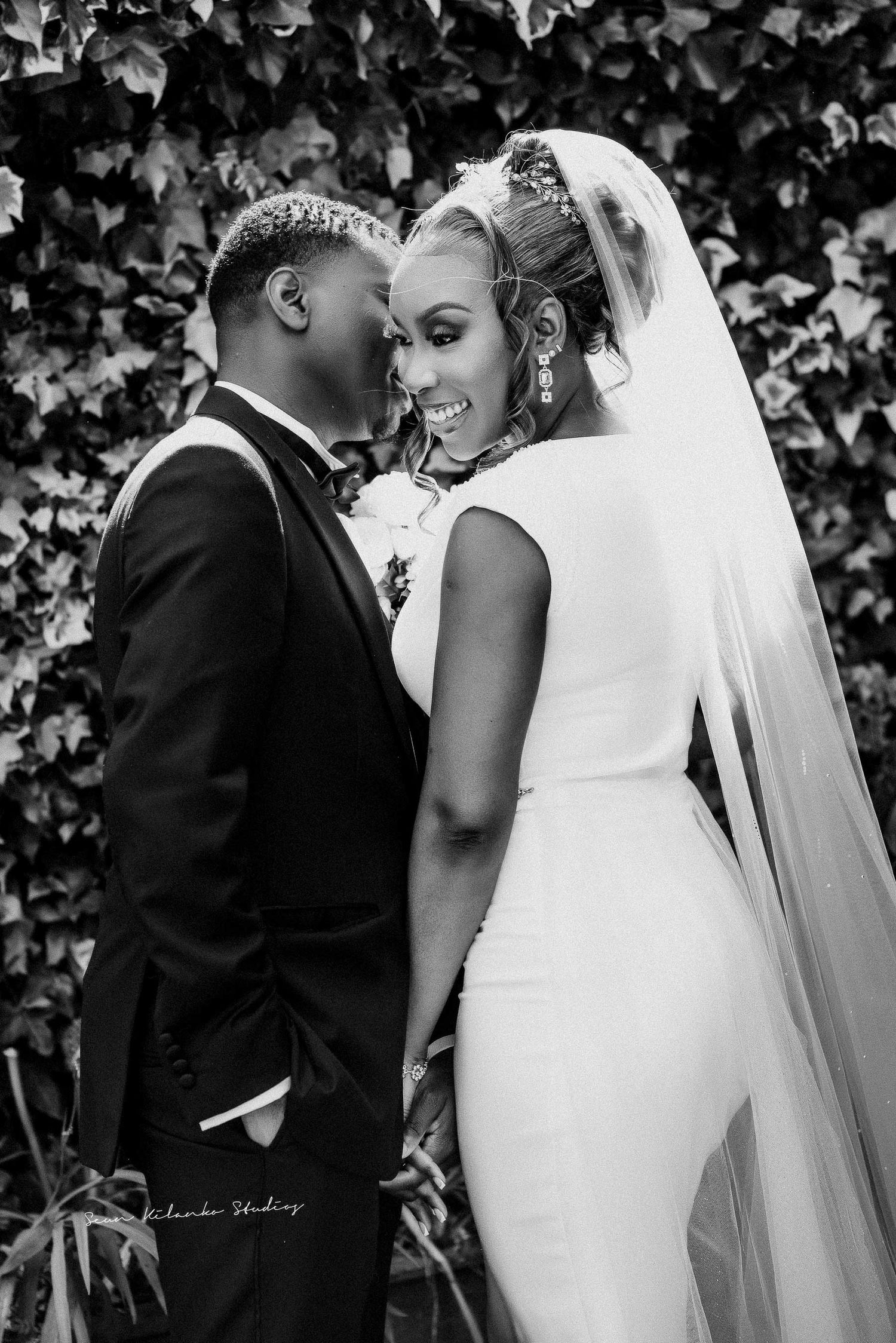 Black and white portrait of newlyweds in an intimate pose against an ivy wall.
