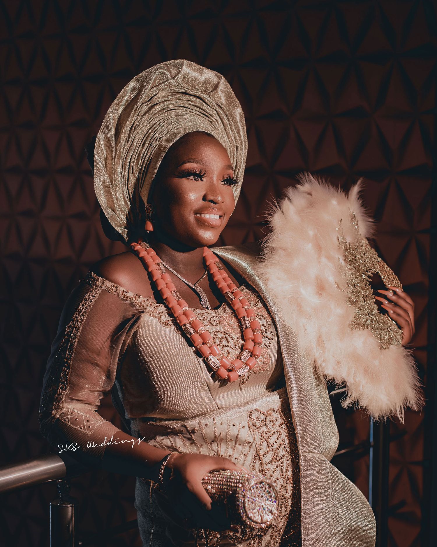 A figure in traditional Nigerian attire with a gold headwrap, beaded necklaces, and sheer dress holding an ornate feather fan.