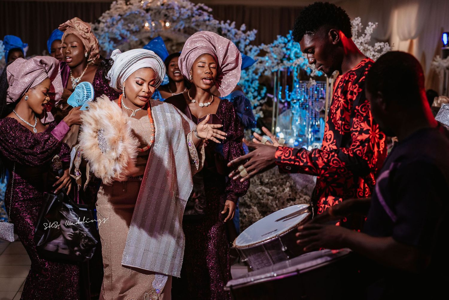 A group of wedding guests in headscarves dancing and celebrating joyfully at an indoor reception with blue decor.