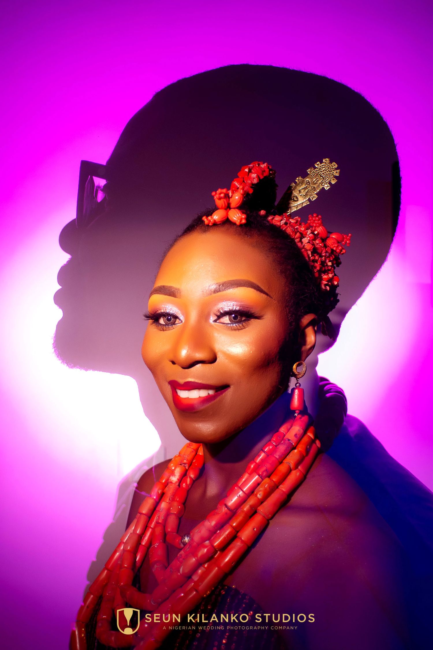 Portrait of figure wearing red beaded necklaces and ornate headpiece against vibrant pink background with dramatic lighting.