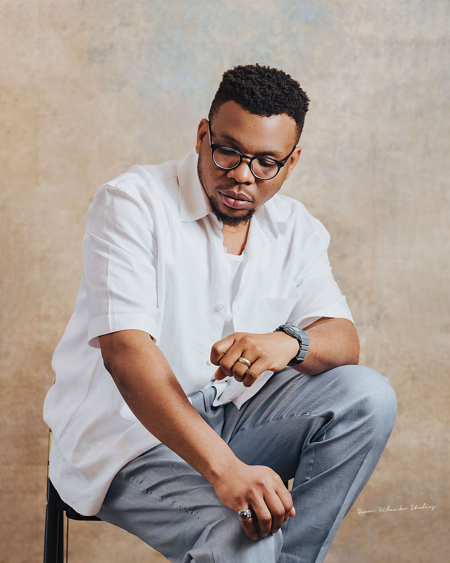 Contemplative studio portrait of a person in white shirt and gray pants seated against textured backdrop.