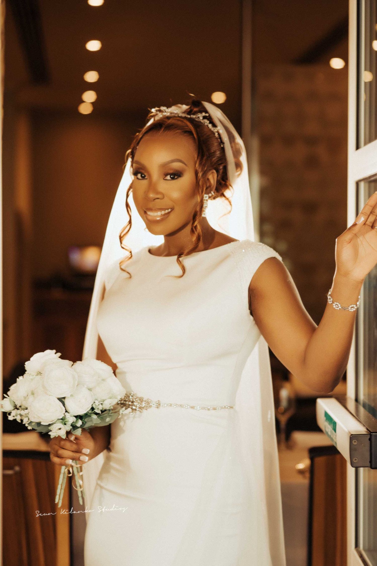 A bride in a classic white dress holds a bouquet of white roses while standing near an indoor doorway.