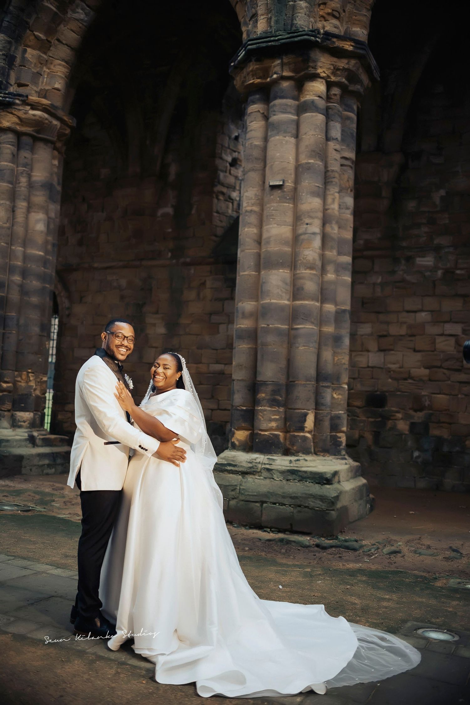 A wedding couple poses beneath grand stone archways of a historic cathedral in elegant white attire.