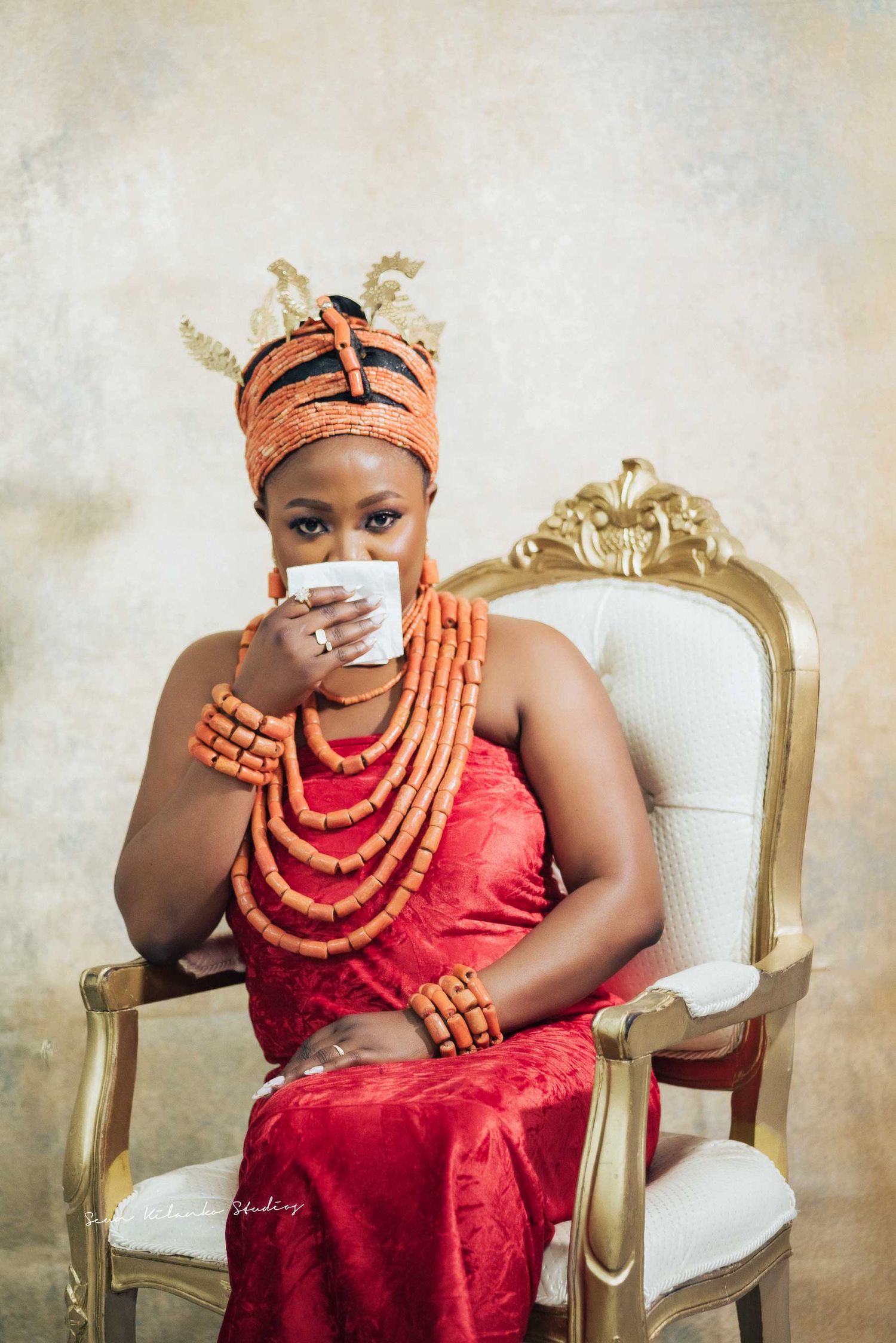 Portrait in red dress and coral beads holding white teacup while seated in gold chair.