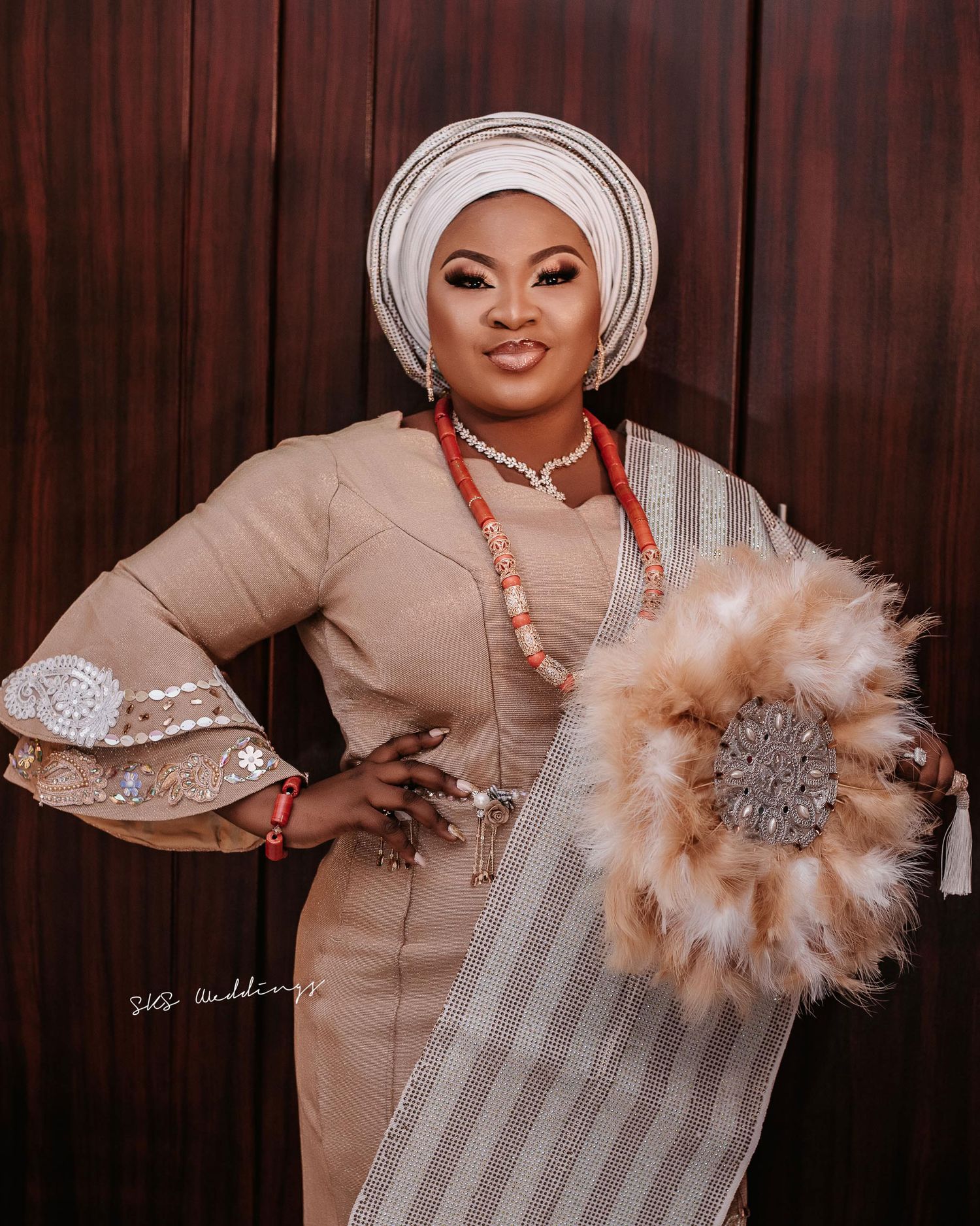 Woman in beige fitted dress with bell sleeves and traditional white head wrap poses elegantly with feathered and beaded accessories.