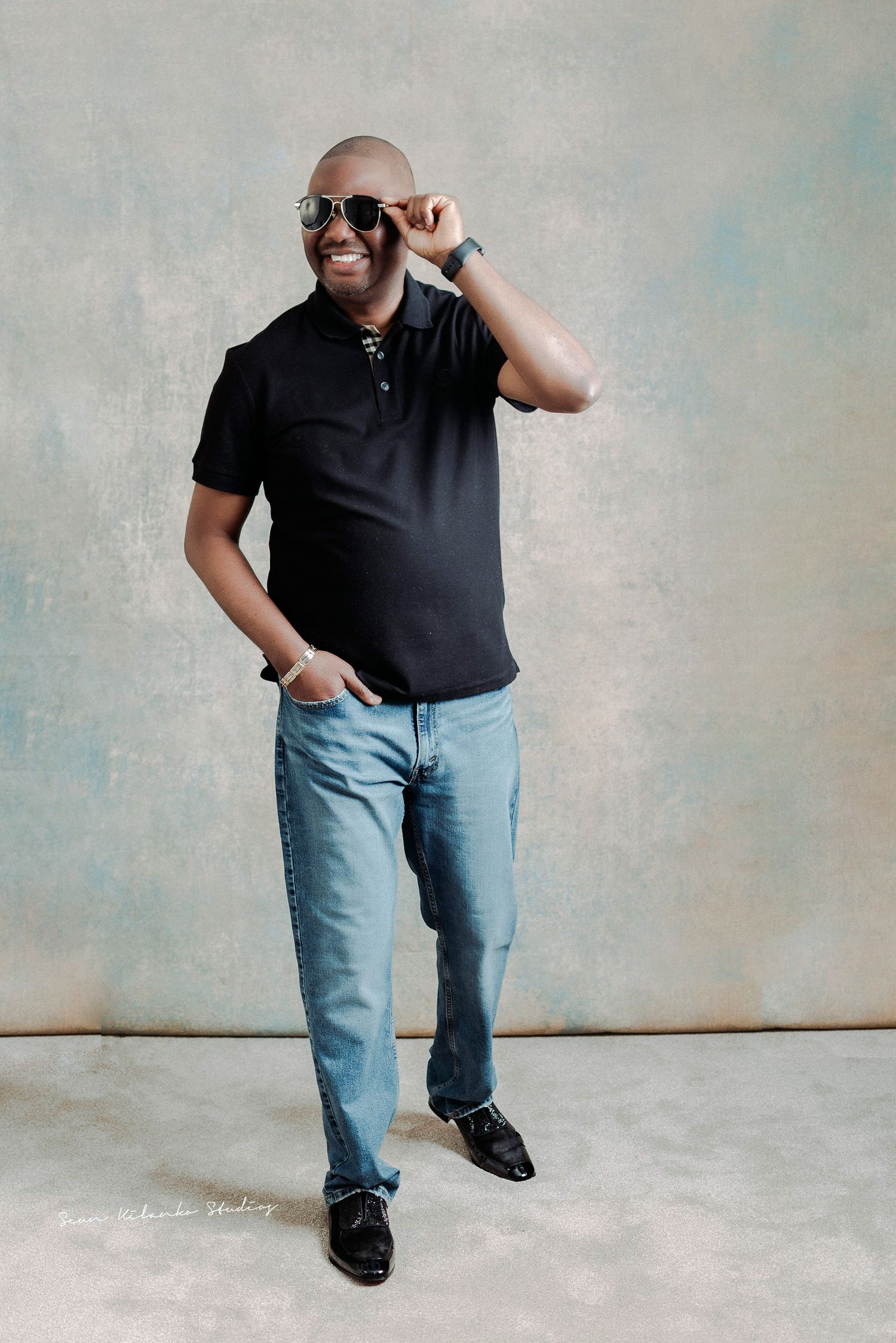 Casual portrait of someone in black polo and jeans with sunglasses against light background.