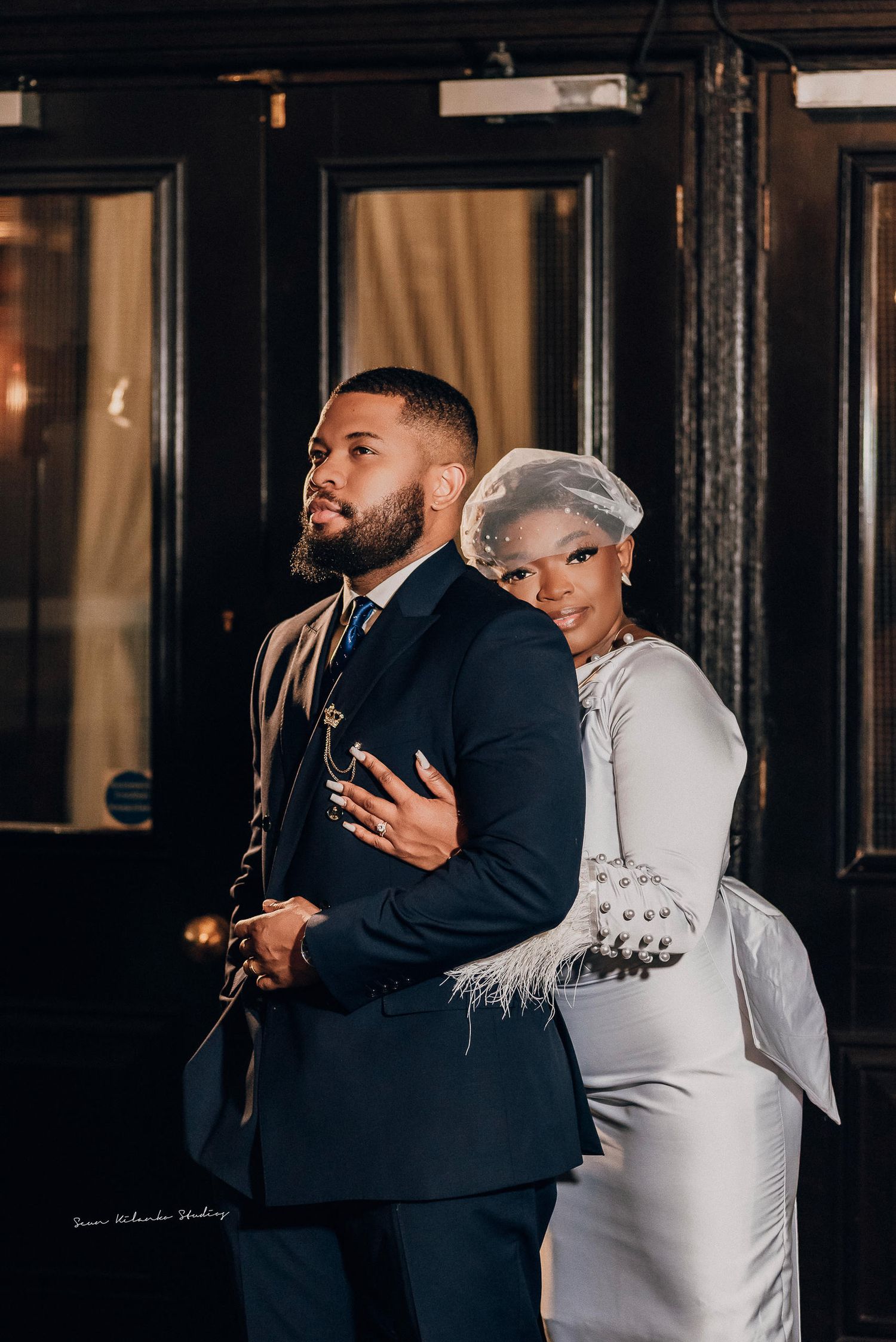 Elegant couple posing in formal attire, man in navy suit and woman in white gown with feather accents at upscale venue.