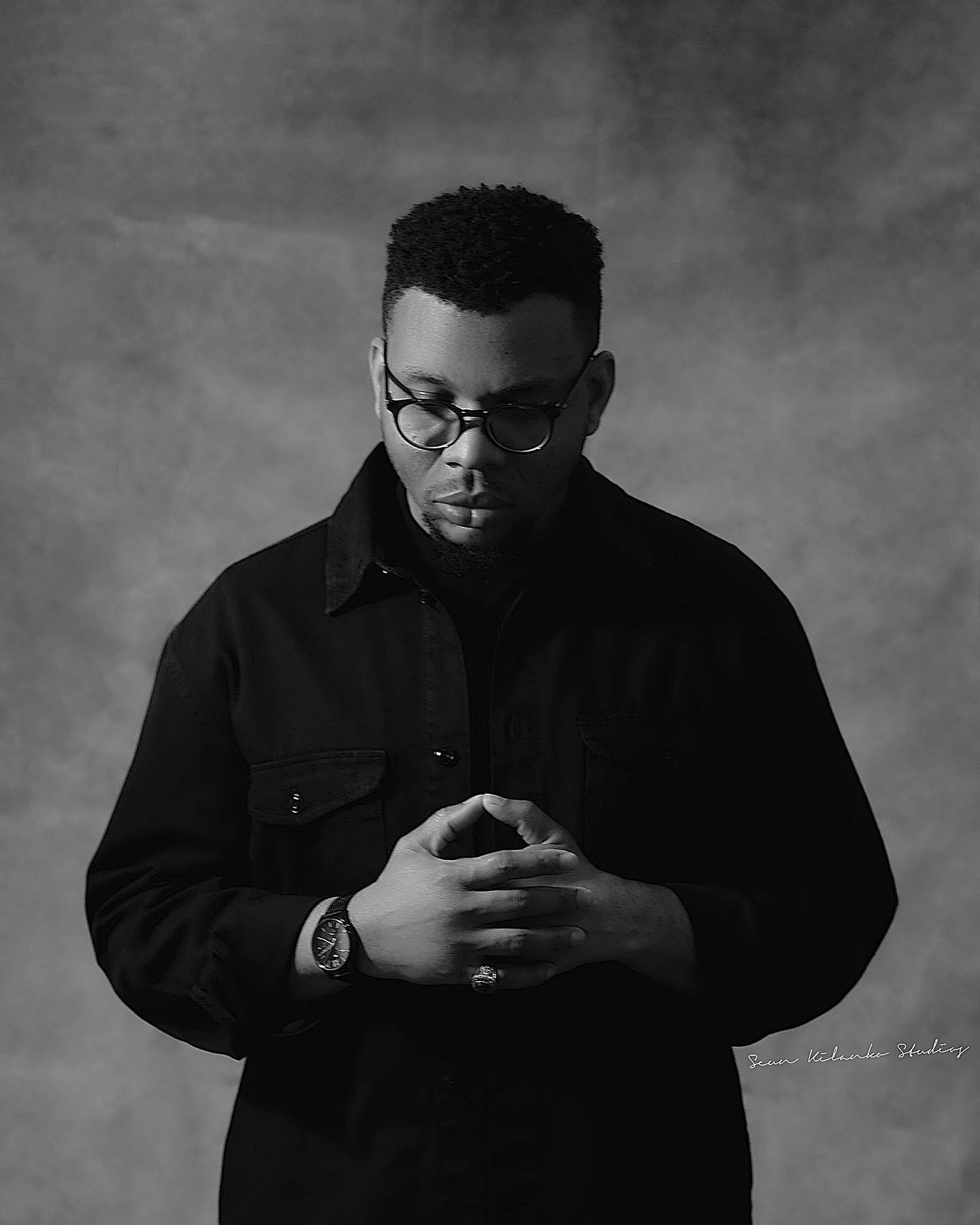 Dramatic black and white portrait of a person in dark clothing with downward gaze.