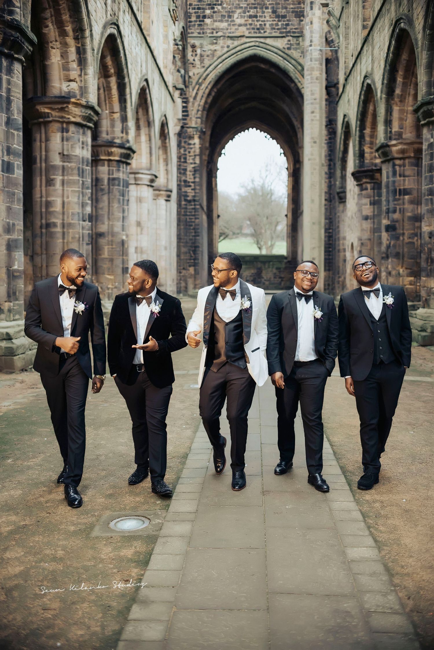 Groomsmen in tuxedos walk together through a gothic cathedral hallway.