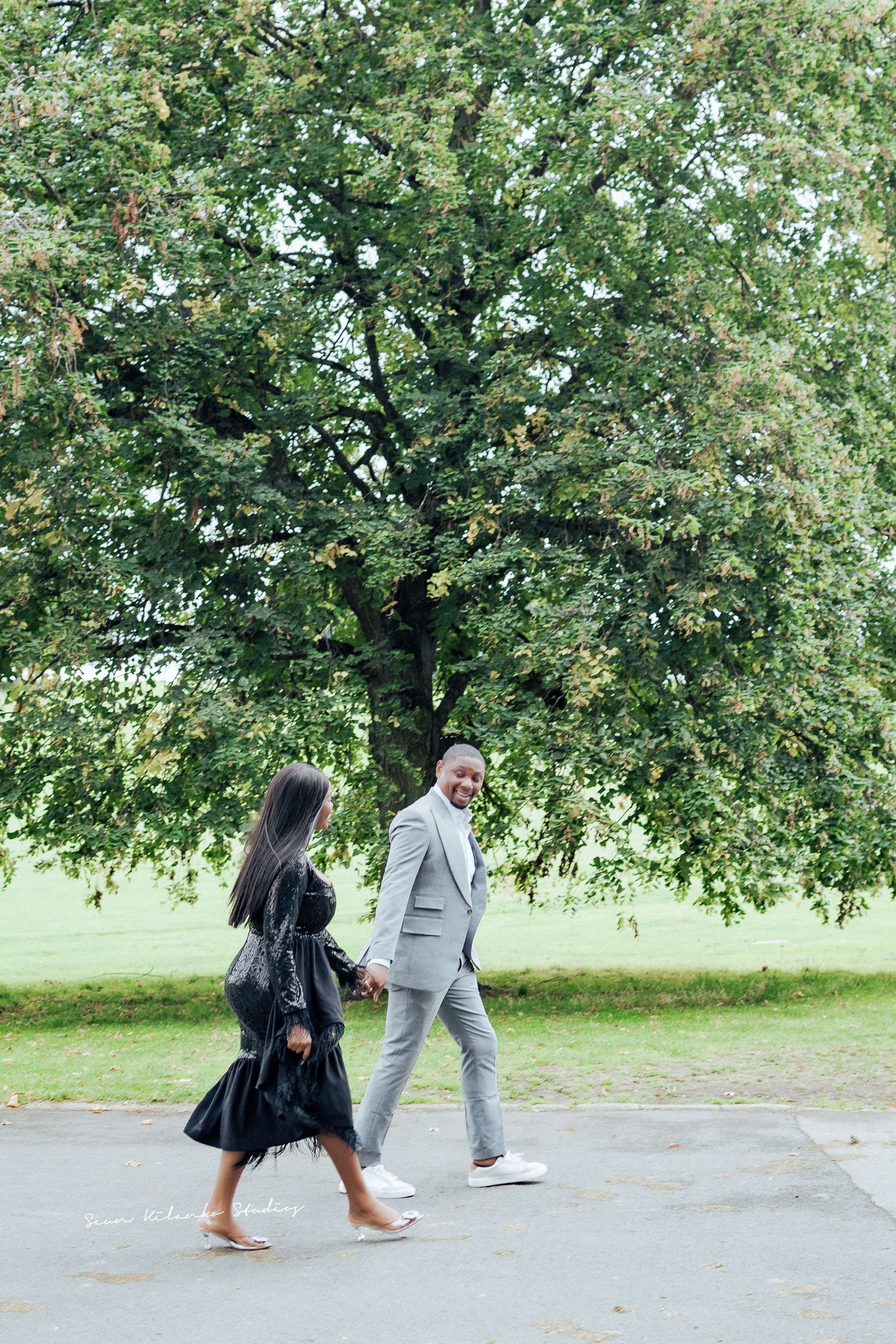 Couple in formal wear walking beneath a large tree in a park setting.