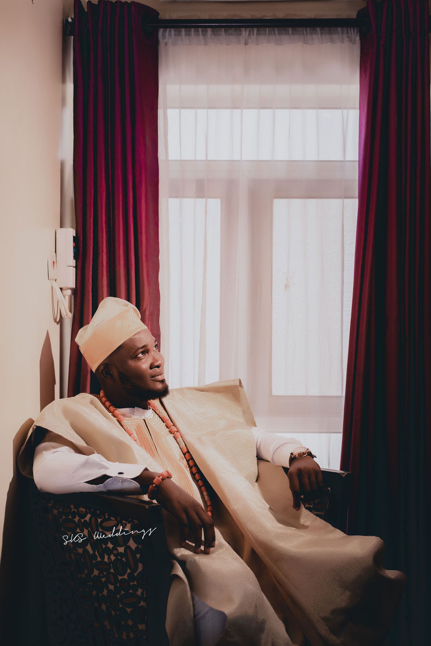 Person lounging on cream-colored couch wearing light-colored outfit and cap near burgundy curtains and window.