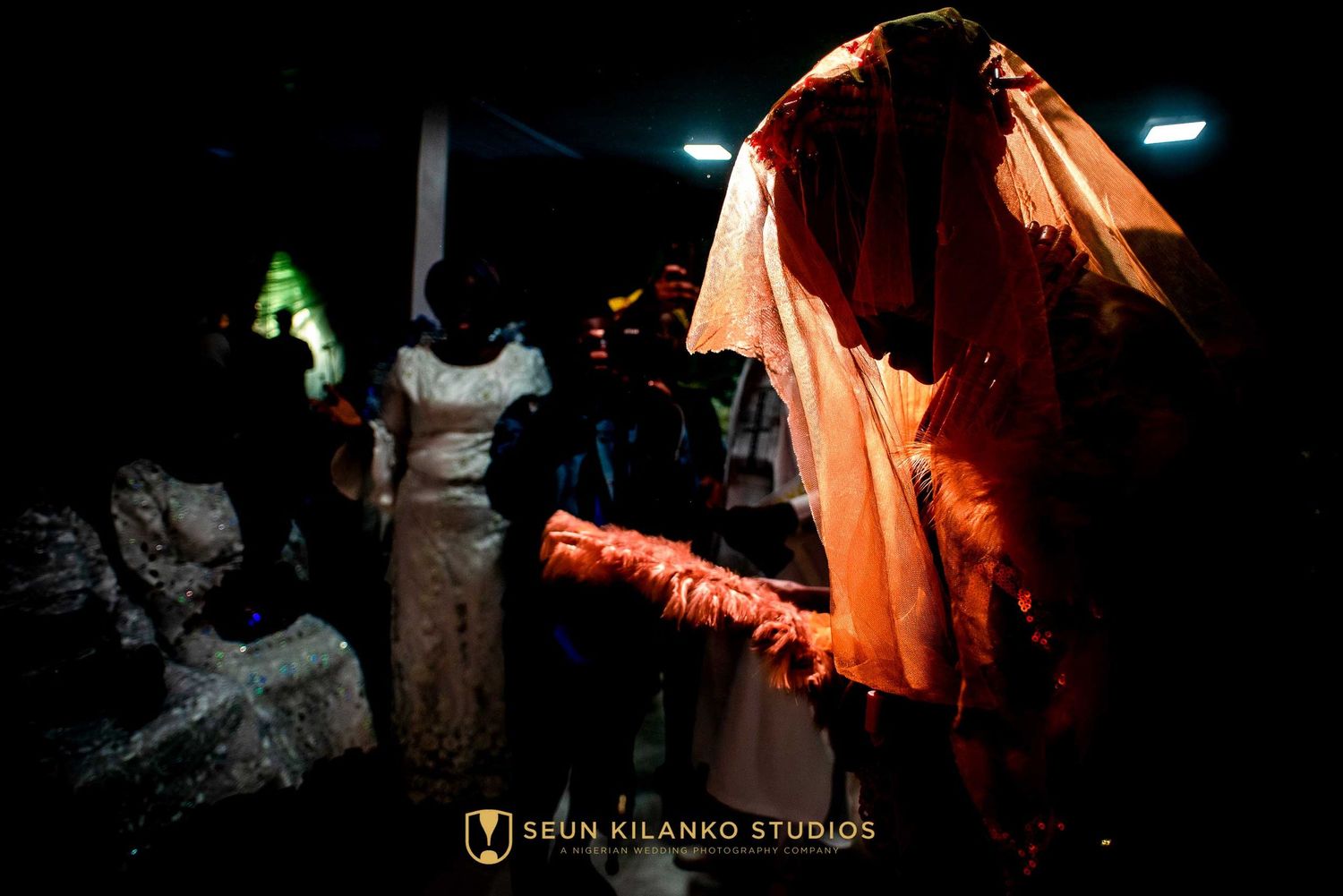 A figure in a glowing orange dress illuminated in darkness during what appears to be a nighttime celebration.