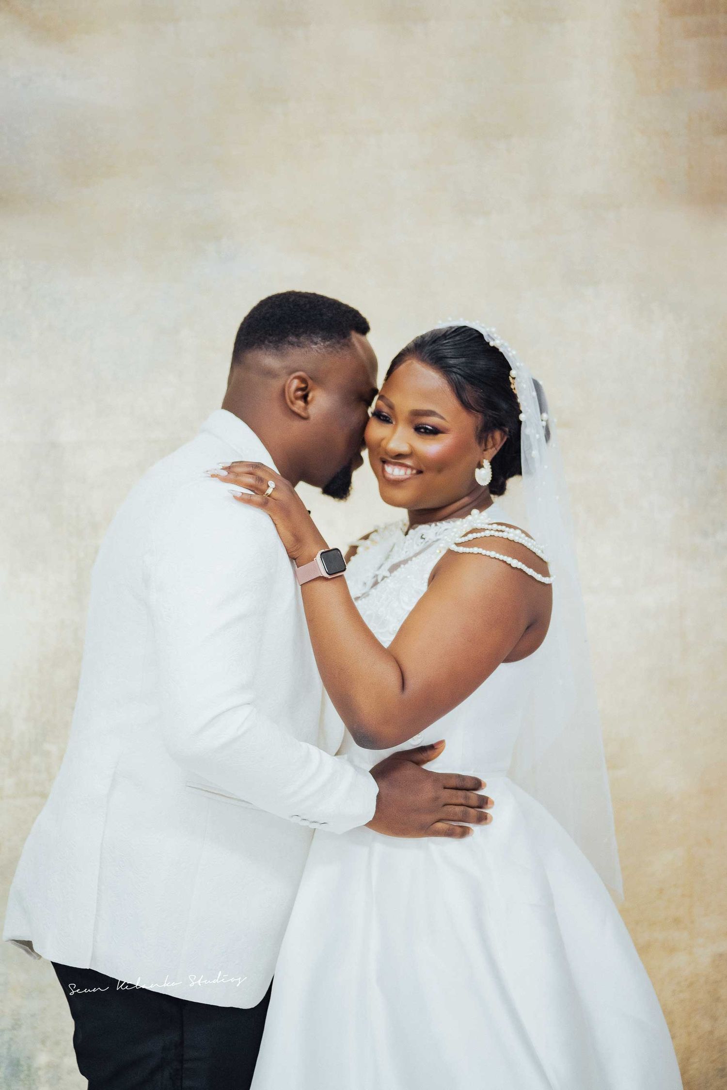 A romantic wedding portrait of a couple in white attire embracing against a neutral background.