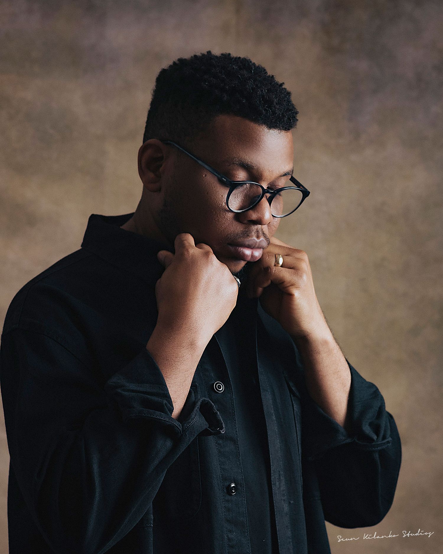 Artistic portrait of a person in dark clothing touching face thoughtfully against warm backdrop.
