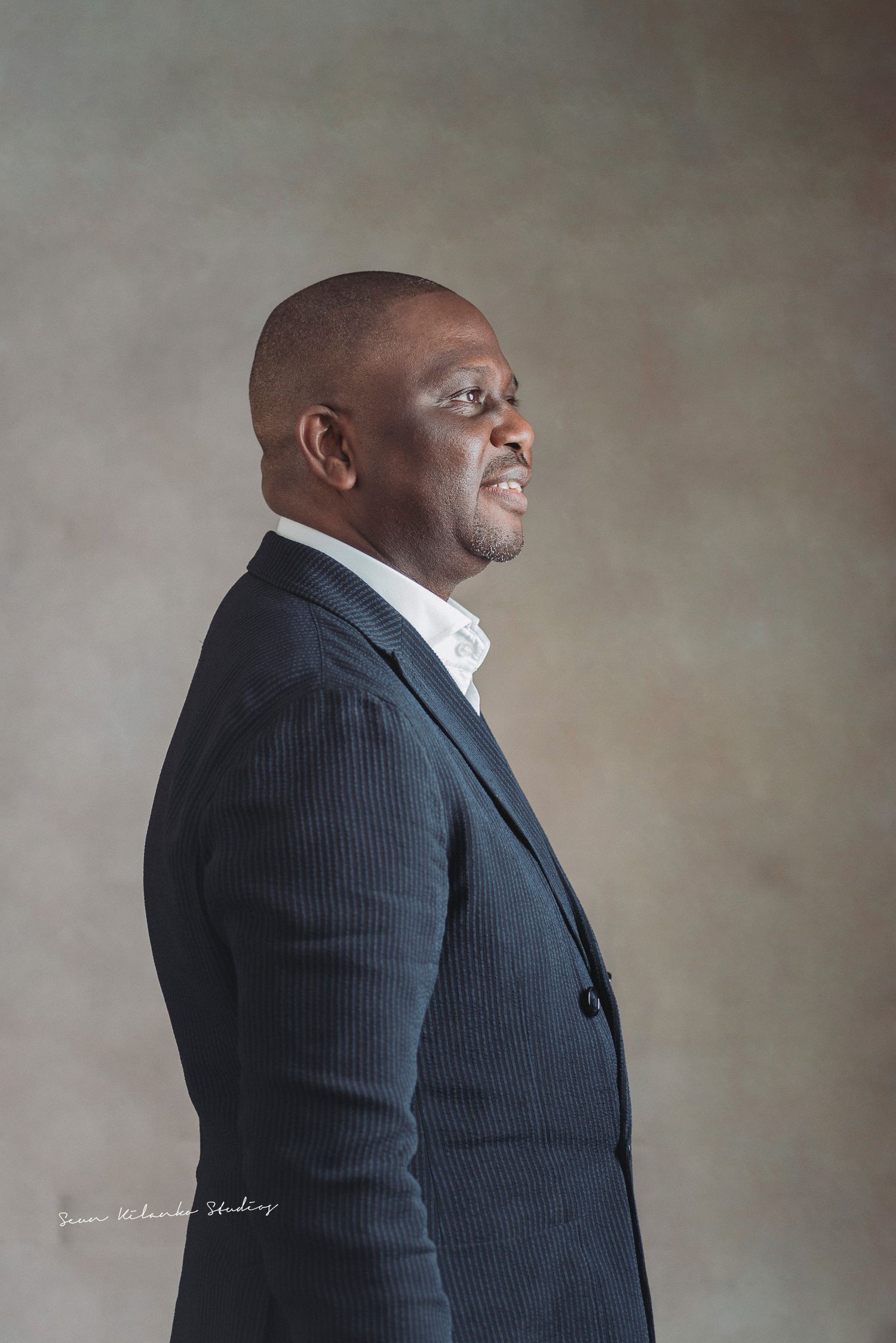 Side profile studio portrait of someone in pinstriped suit jacket against neutral background.