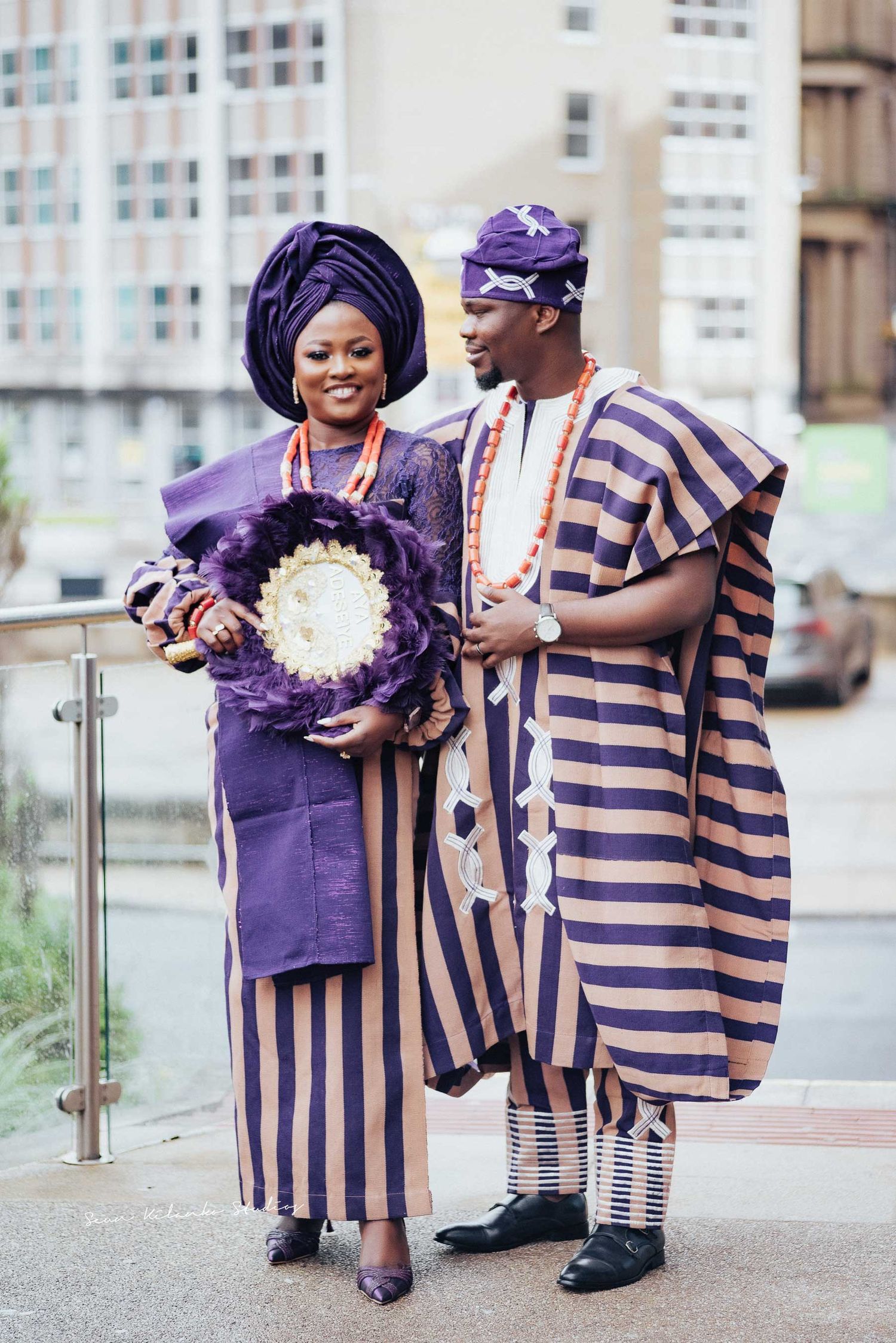 Couple in coordinated Nigerian attire smiling while holding decorative white bouquet.