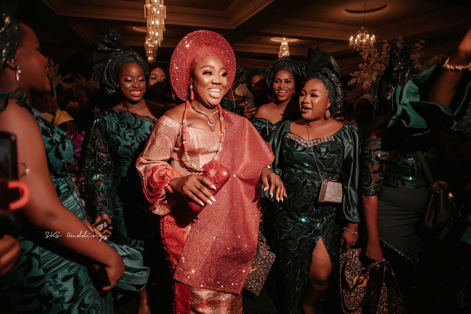 Party guests in traditional Nigerian attire dance and celebrate at an elegant event with dim red lighting.