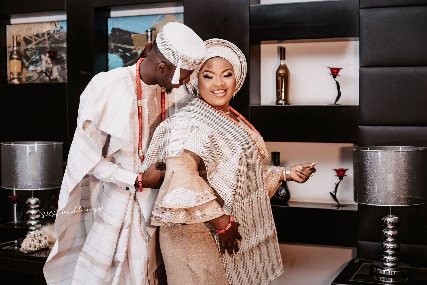 A couple in elegant white traditional Nigerian wedding attire dancing together in a stylishly decorated indoor setting.