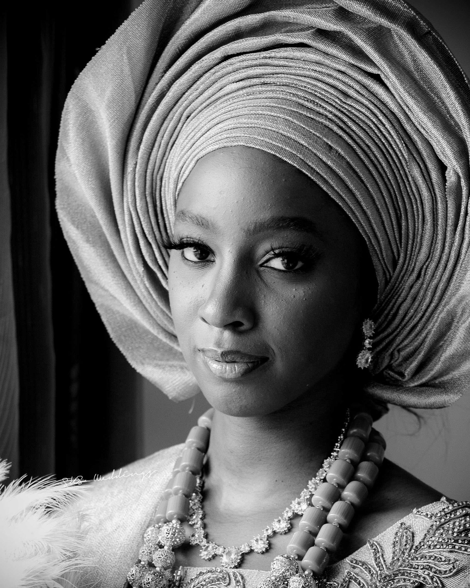 Black and white portrait of an elegant woman wearing a traditional African head wrap, beaded necklace and lace attire.